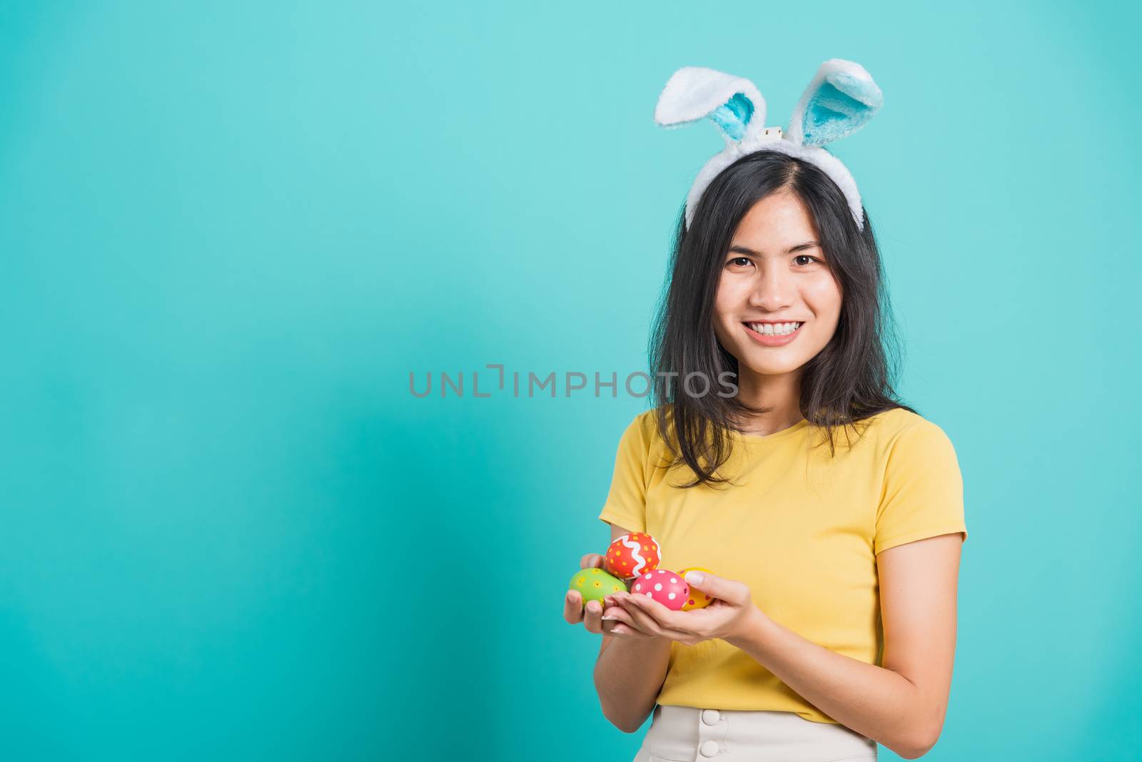 Portrait Asian beautiful happy young woman smile white teeth wear yellow t-shirt standing with bunny ears and holding Easter eggs looking to camera, on a blue background with copy space