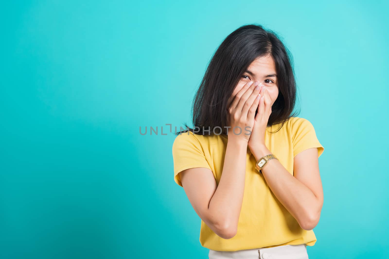 Portrait Asian beautiful happy young woman wears yellow t-shirt she happy excited laughs covering mouth with hands, shoot photo in a studio, on a blue background, with copy space