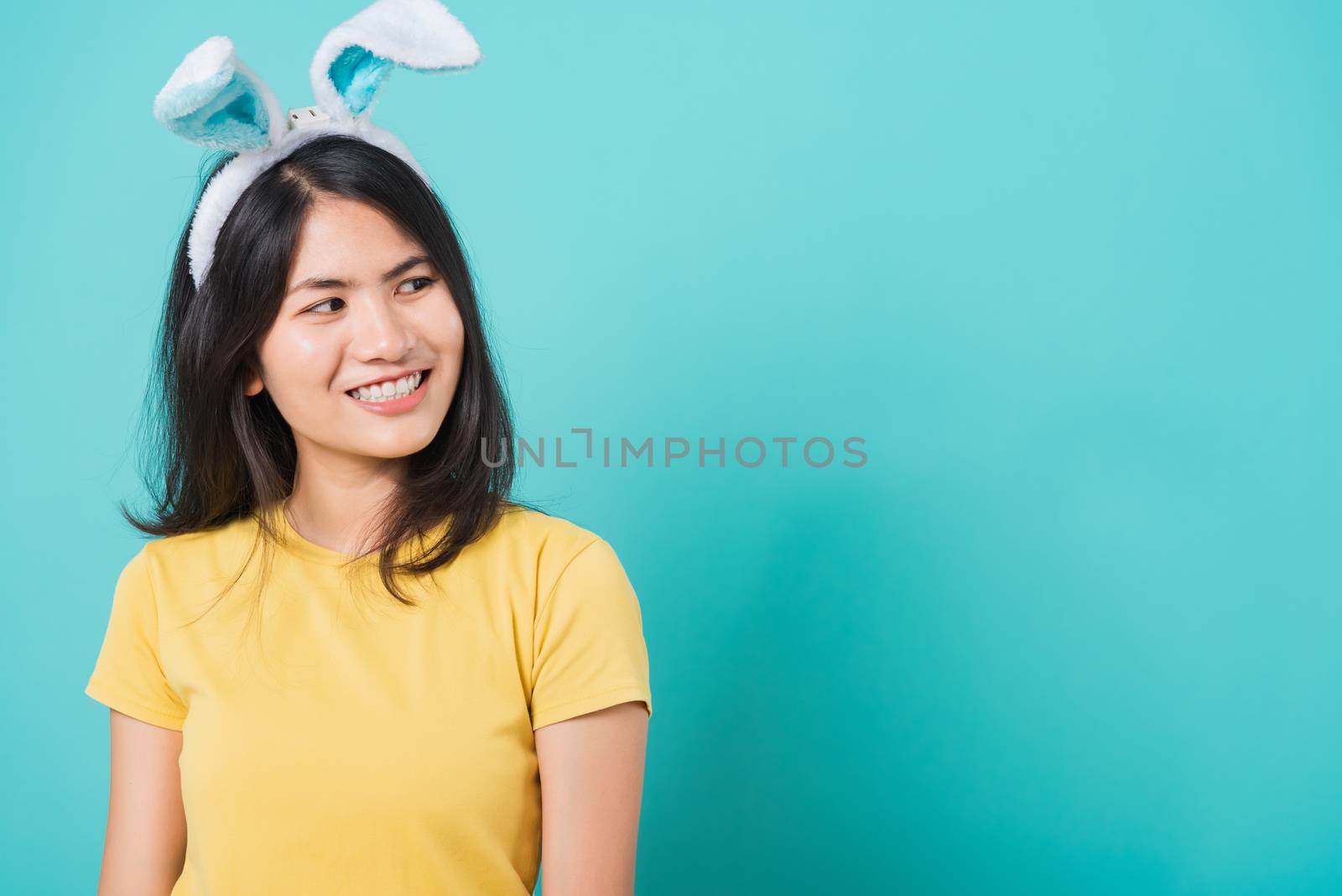 Portrait Asian beautiful happy young woman smile white teeth wear yellow t-shirt standing with bunny ears her looking to space, on blue background with copy space