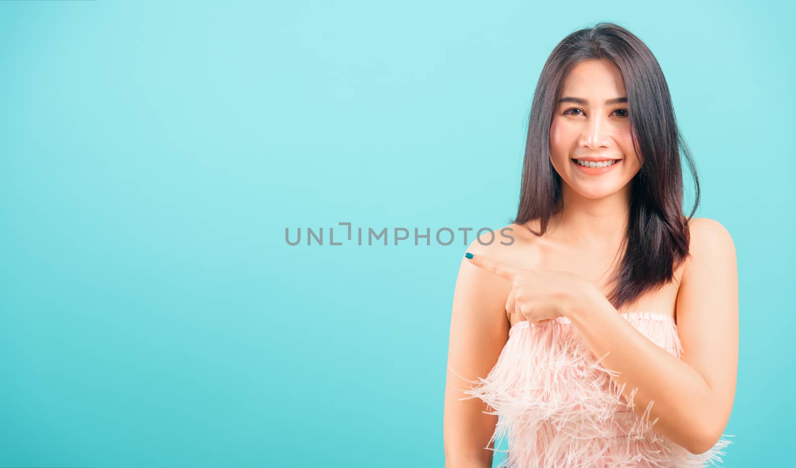 Smiling face Asian beautiful woman her point finger beside and looking to camera on blue background, with copy space for text