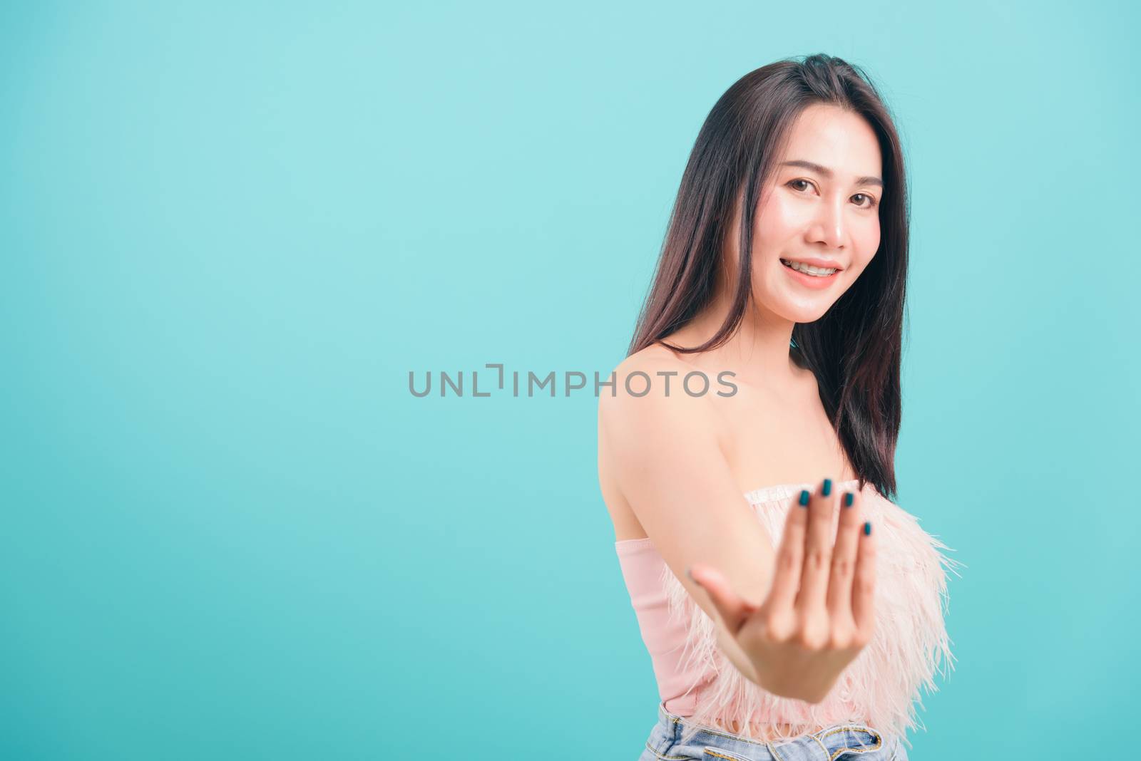 Asian happy portrait beautiful young woman standing smiling gesture positive with hand inviting to come here sign and looking to camera on blue background with copy space for text