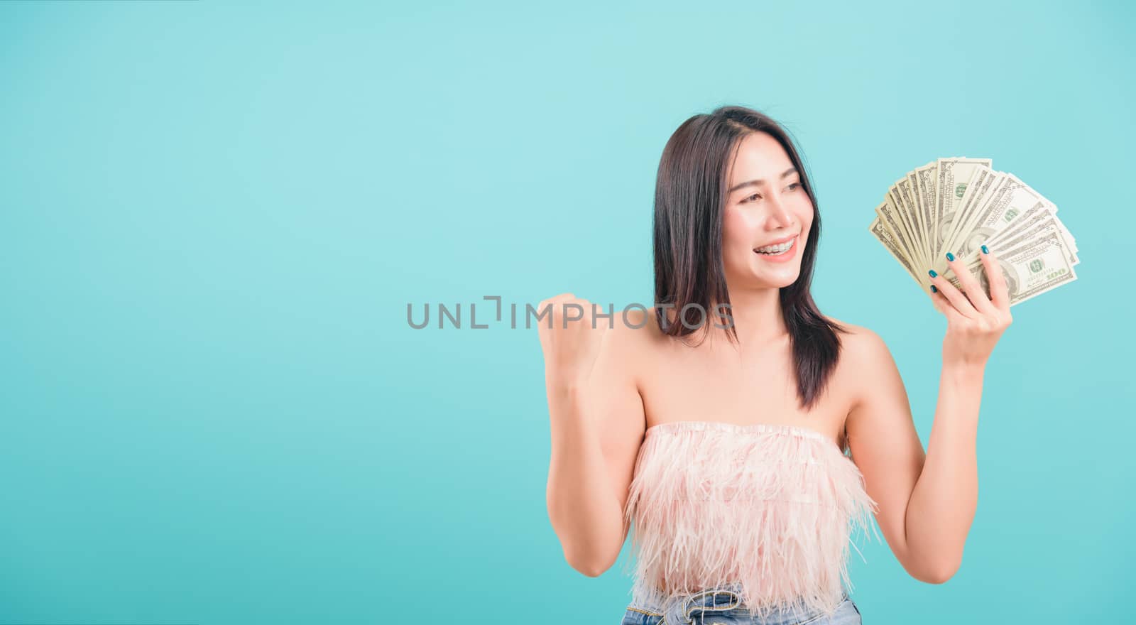 Asian happy portrait beautiful young woman standing smile her celebrating holding dollar money fan banknotes on hand and looking to camera on blue background with copy space for text