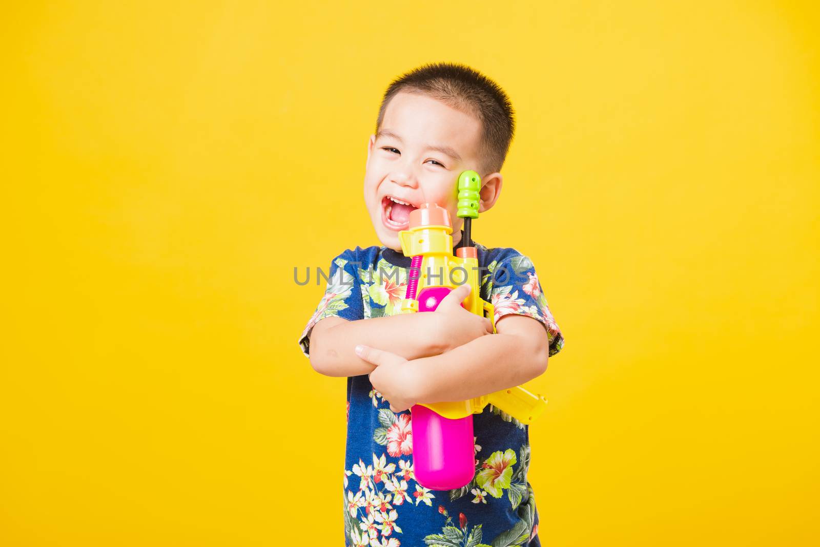 Portrait happy Asian cute little children boy smile standing so happy wearing flower shirt in Songkran festival day holding water gun, studio shot on yellow background with copy space