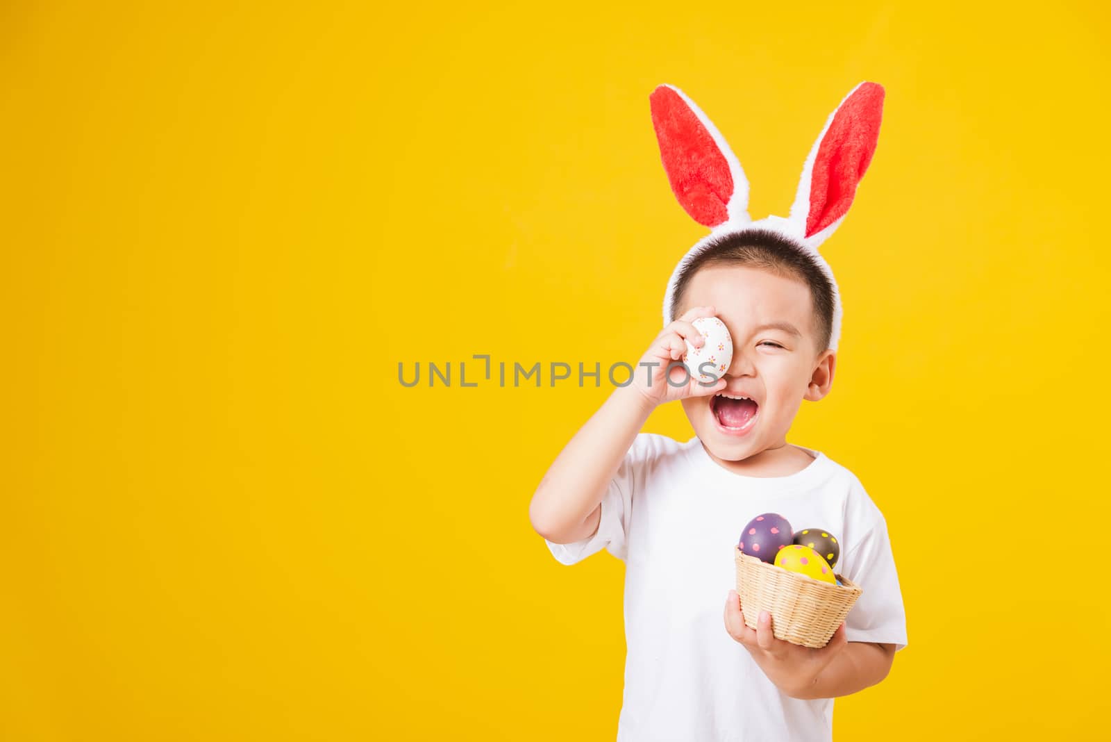 Portrait happy Asian cute little children boy smile standing so happy wearing white T-shirt and bunny ears in Easter festival day holding easter eggs, studio shot on yellow background with copy space