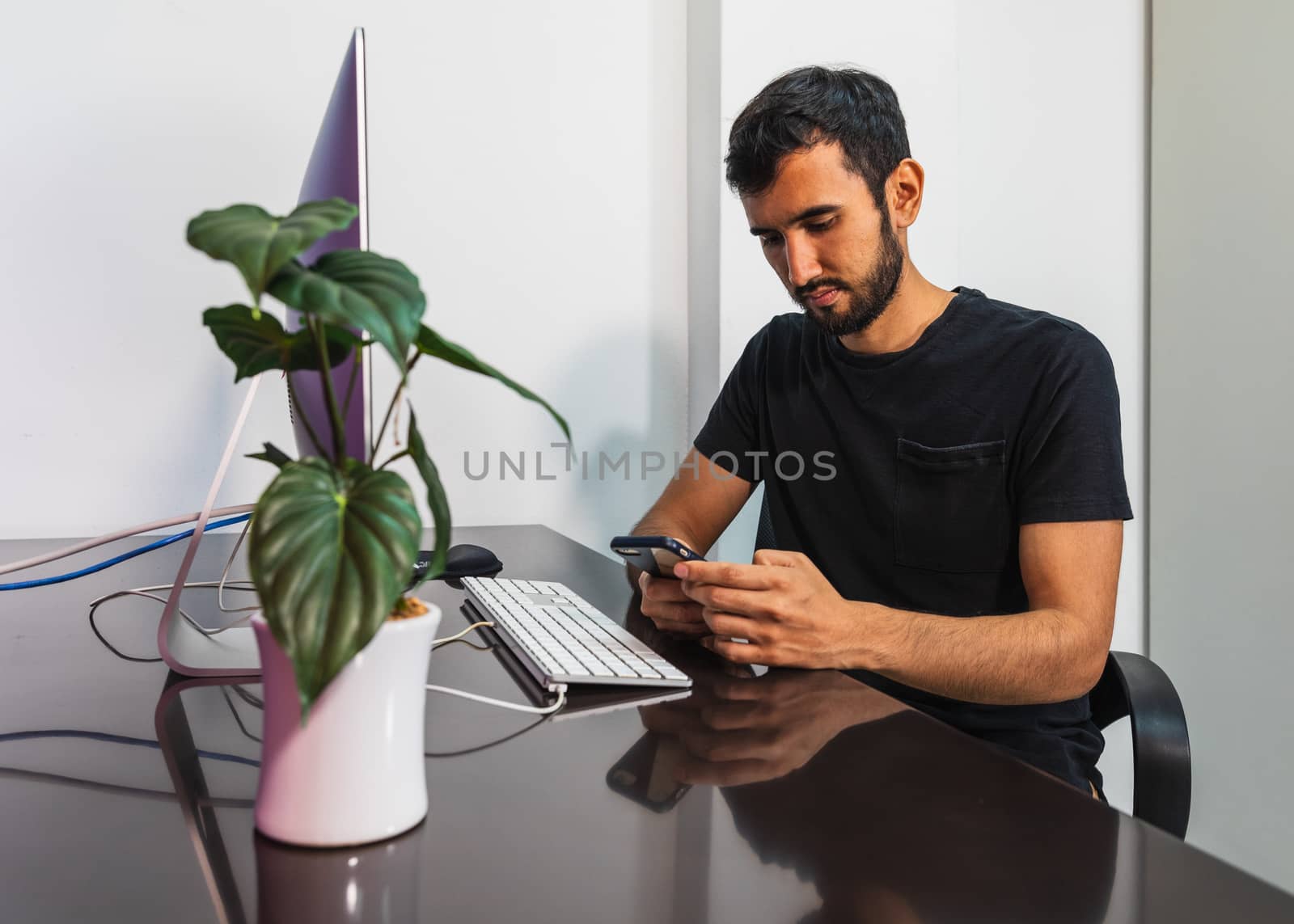Bearded man in a casual wear holding smart phone and looking at it while sitting at his working place in office
