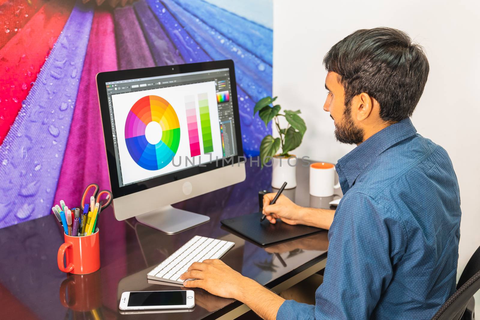 Side view. Young bearded businessman in denim shirt is sitting in office at table and is using computer with color swatch on screen. On table is smartphone and stationery. Man working.