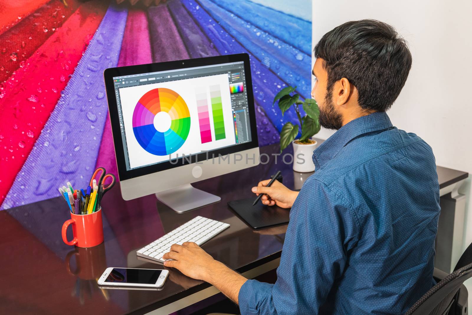 Side view. Young bearded businessman in denim shirt is sitting in office at table and is using computer with color swatch on screen. On table is smartphone and stationery. Man working.