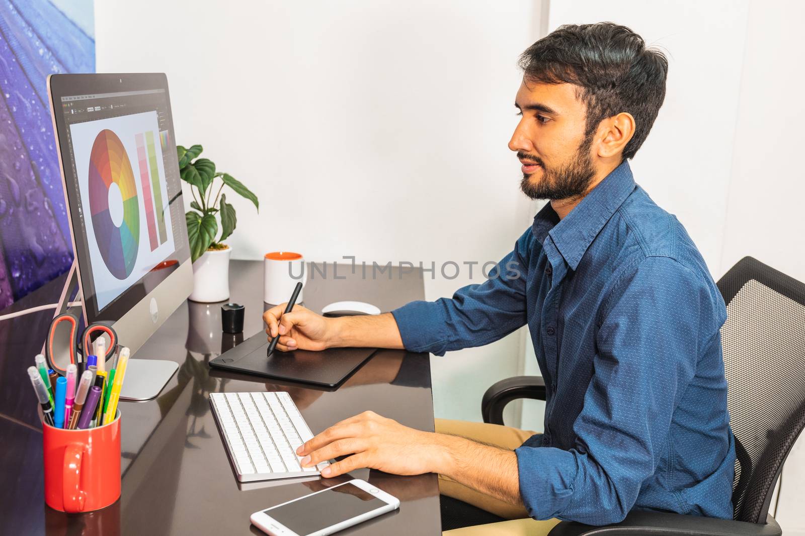 Side view. Young bearded businessman in denim shirt is sitting in office at table and is using computer with color swatch on screen. On table is smartphone and stationery. Man working