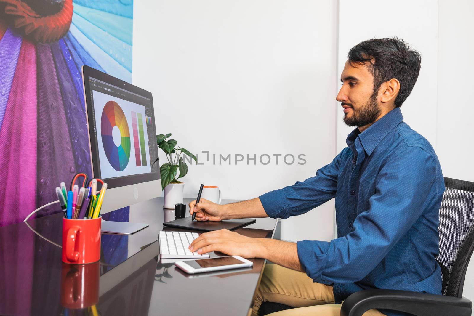 Side view. Young bearded businessman in denim shirt is sitting in office at table and is using computer with color swatch on screen. On table is smartphone and stationery. Man working