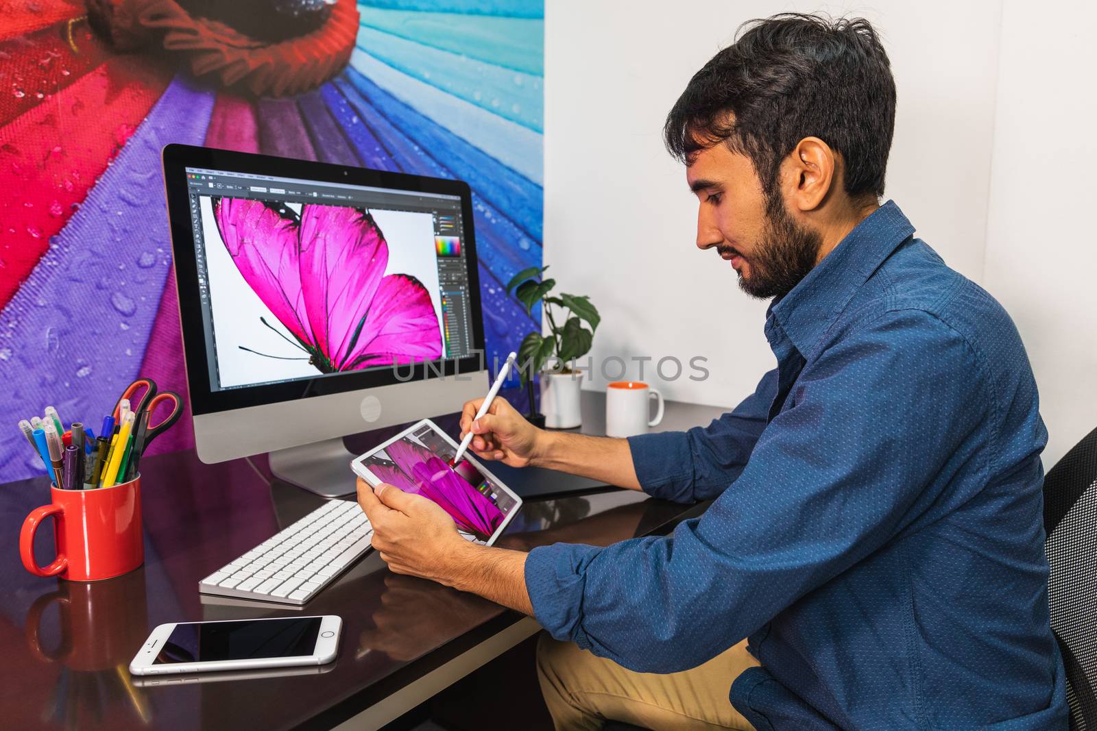 Side view. Young bearded businessman in denim shirt is sitting in office at table and is using a tablet. On table is smartphone and stationery. Man working