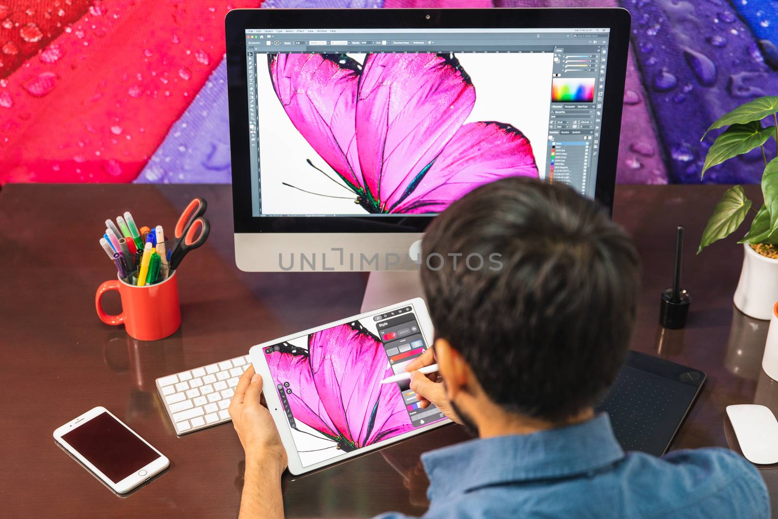 Back view. Young bearded businessman in denim shirt is sitting in office at table and is using a tablet. On table is smartphone and stationery. Man working