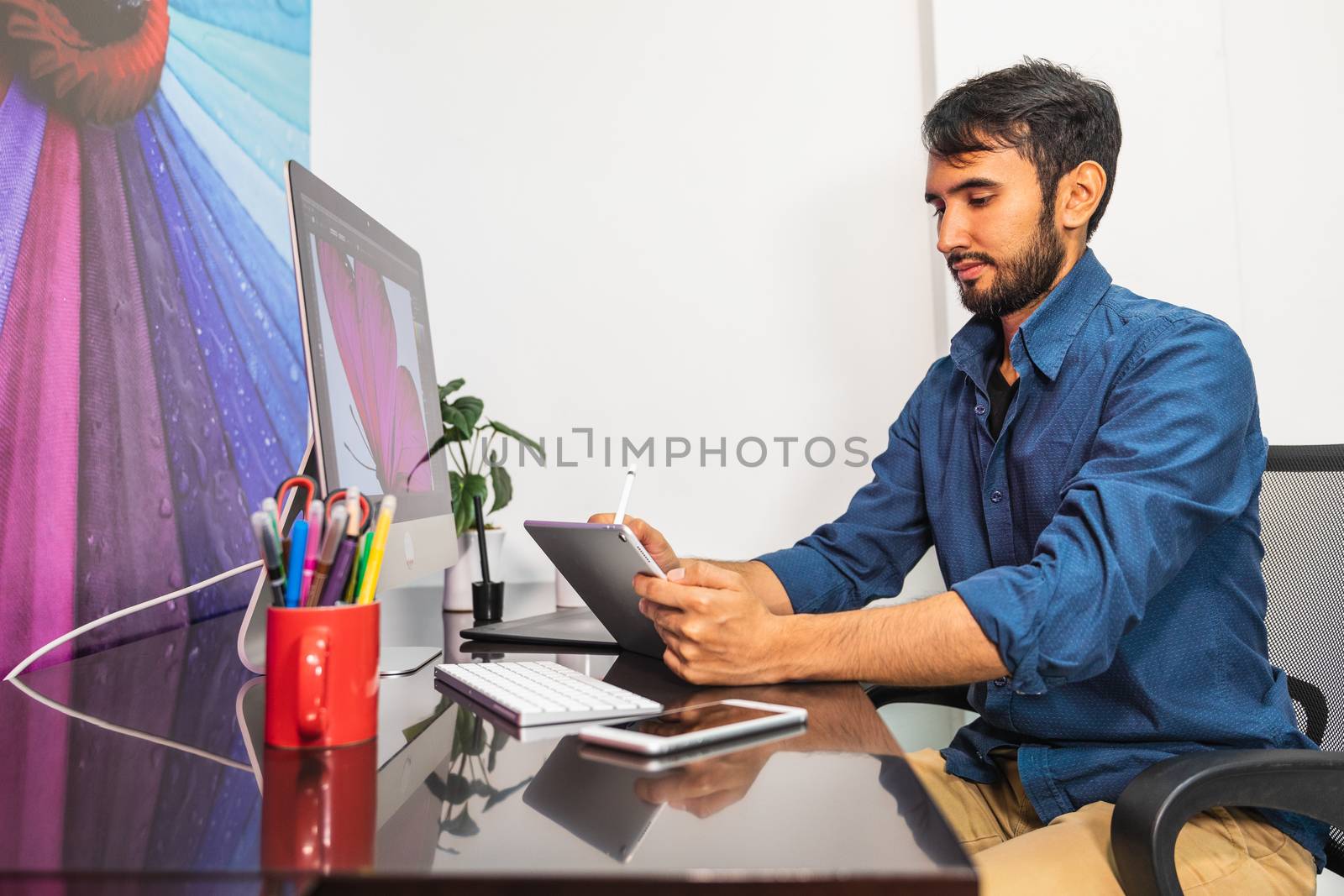 Side view. Young bearded businessman in denim shirt is sitting in office at table and is using a tablet. On table is smartphone and stationery. Man working
