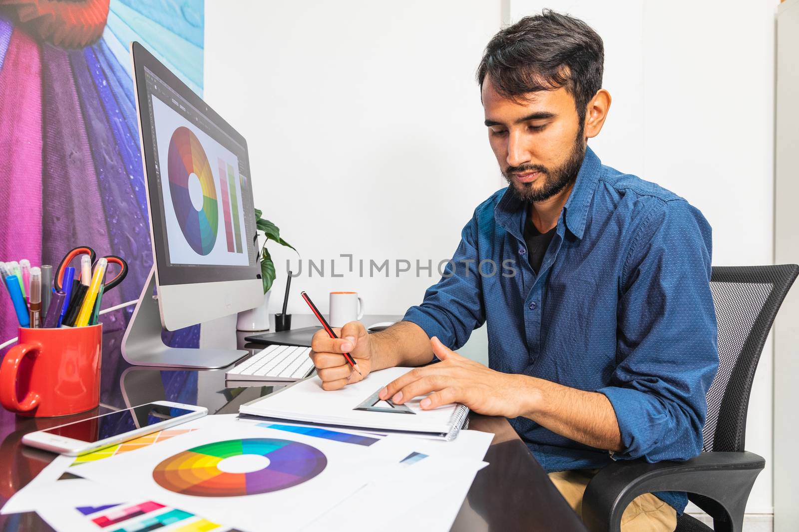 Side view. Young bearded businessman in denim shirt is sitting in office at table. On table is smartphone and stationery. Man drawing sketch