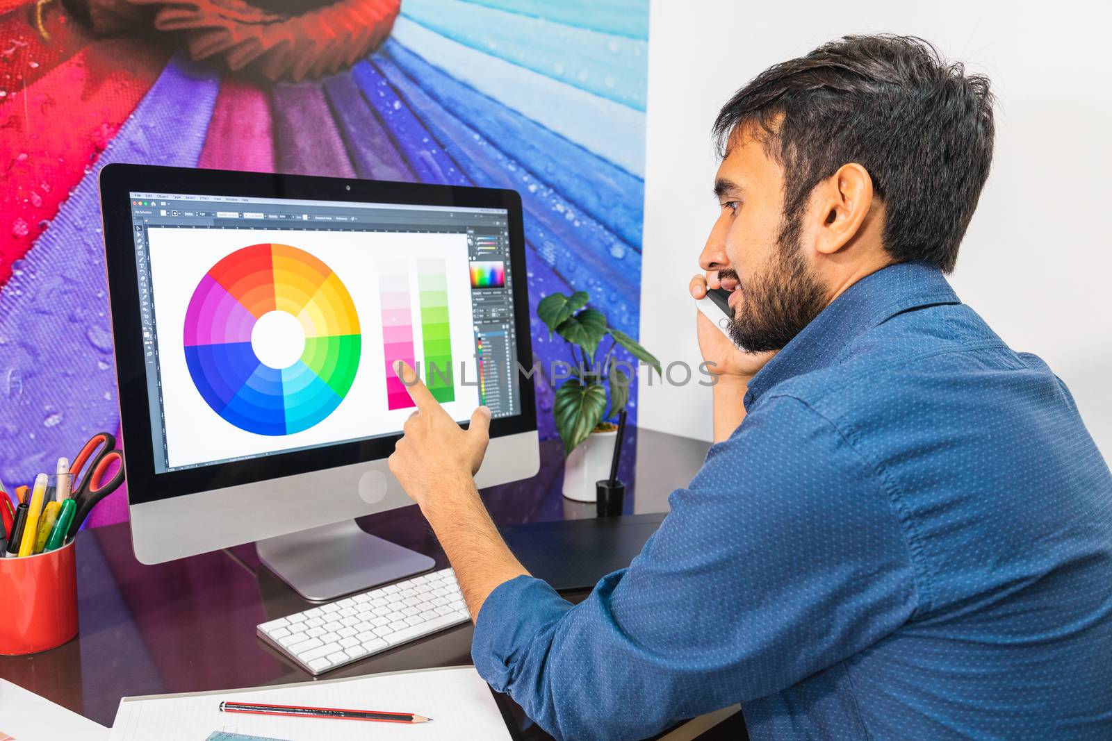 Side view. Young bearded businessman in denim shirt is sitting in office at table. Using his smartphone while looking at color swatch.