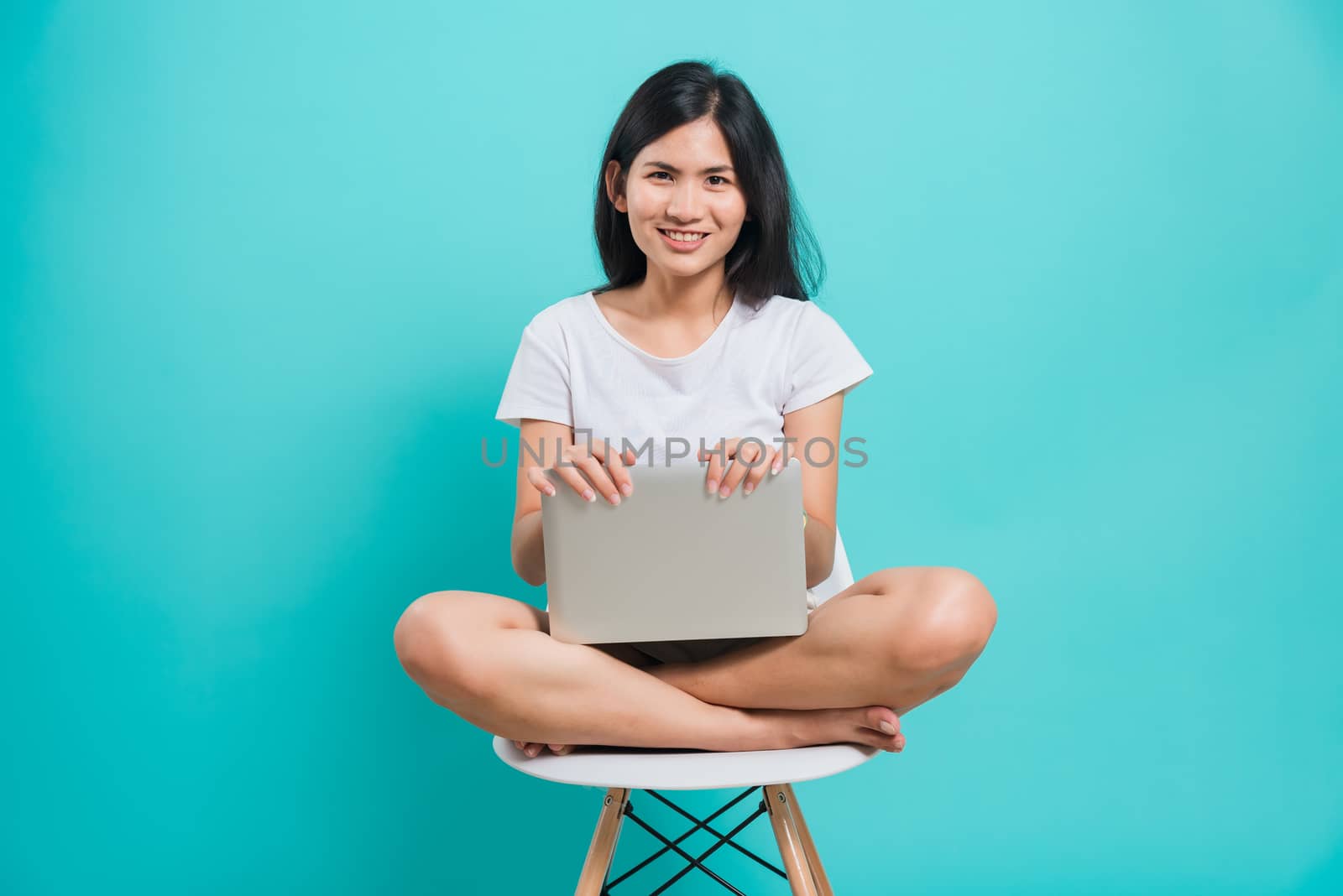 Portrait happy Asian beautiful young woman smile with teeth sitting on chair wear white t-shirt, She holding and using a laptop computer, studio shot on blue background with copy space for text