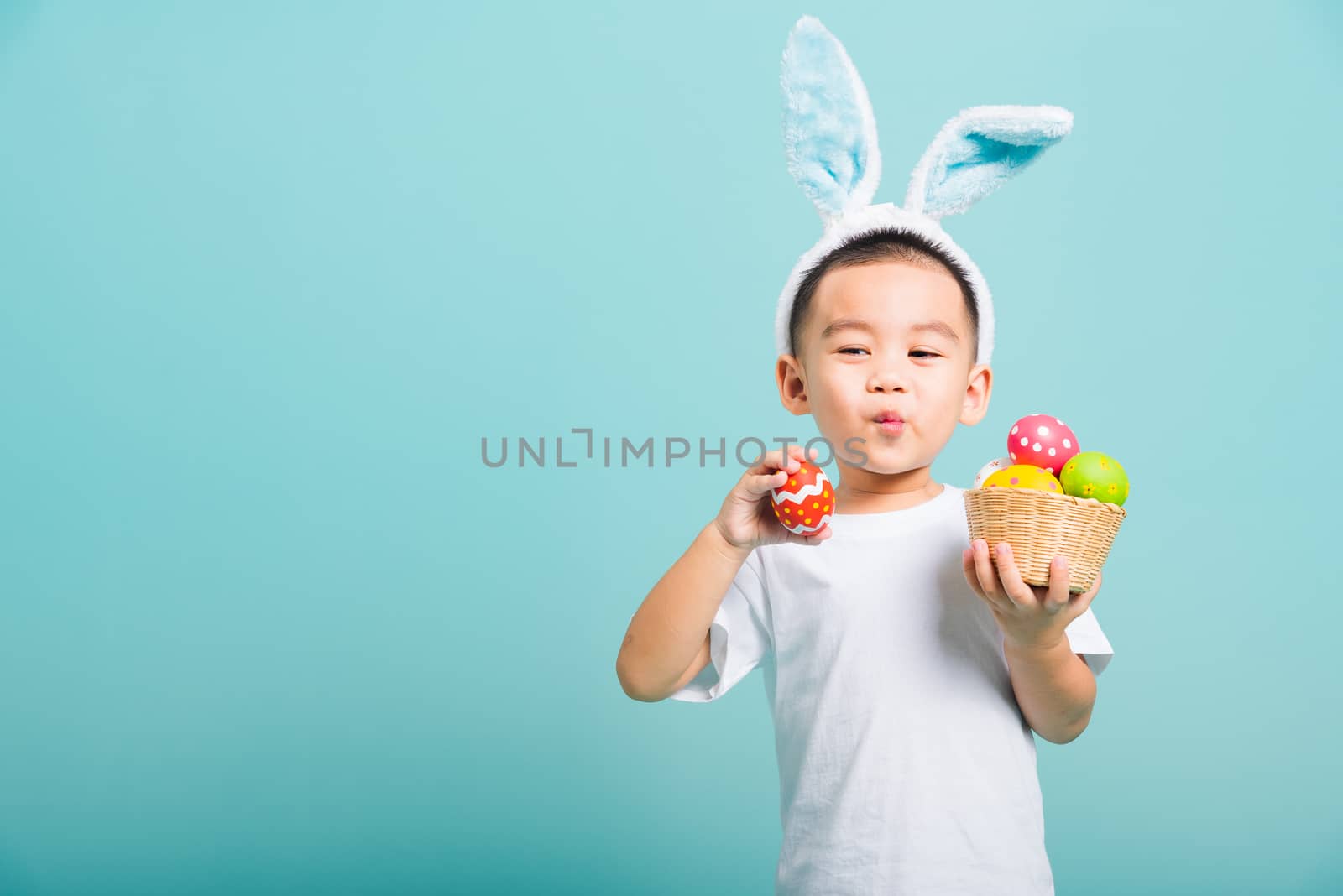 Asian cute little child boy smile beaming wearing bunny ears and a white T-shirt, standing to hold a basket with full Easter eggs. And other hand holds an easter egg on blue background with copy space