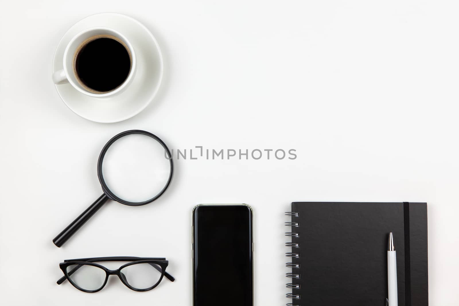 Modern unisex working space, top view. Notepad, pen, coffee, smartphone, magnifier, glasses on white background, copy space, flat lay. Desktop of freelancer. Work from home concept. Monochrome image.