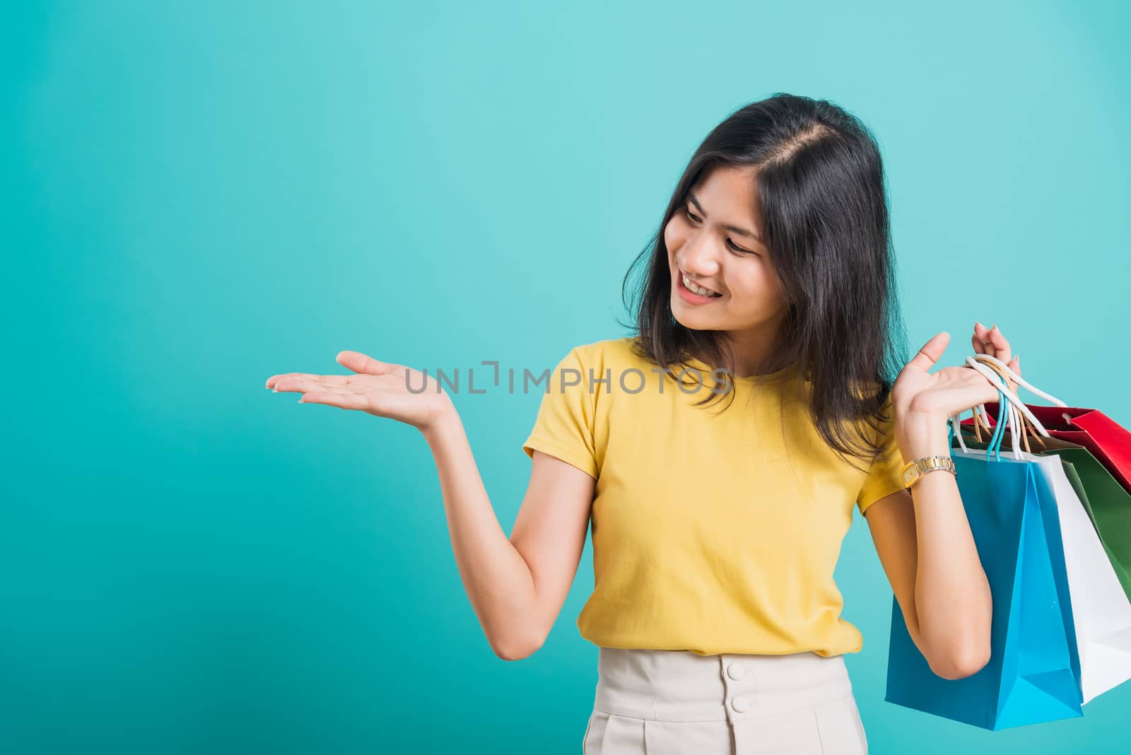 Portrait happy Asian beautiful young woman smile white teeth standing wear yellow t-shirt, She holding shopping bags show hand something empty and looking to space, studio shot on blue background