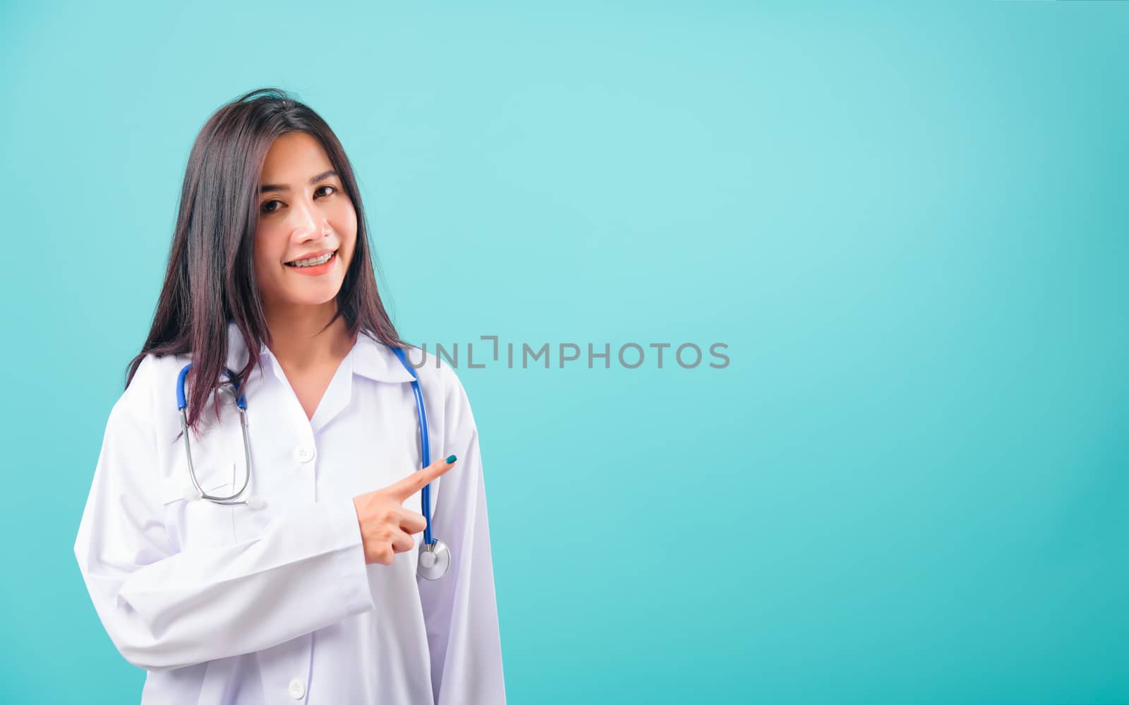 Portrait asian beautiful doctor woman smiling her standing pointing with finger and her looking to camera on blue background, with copy space for text