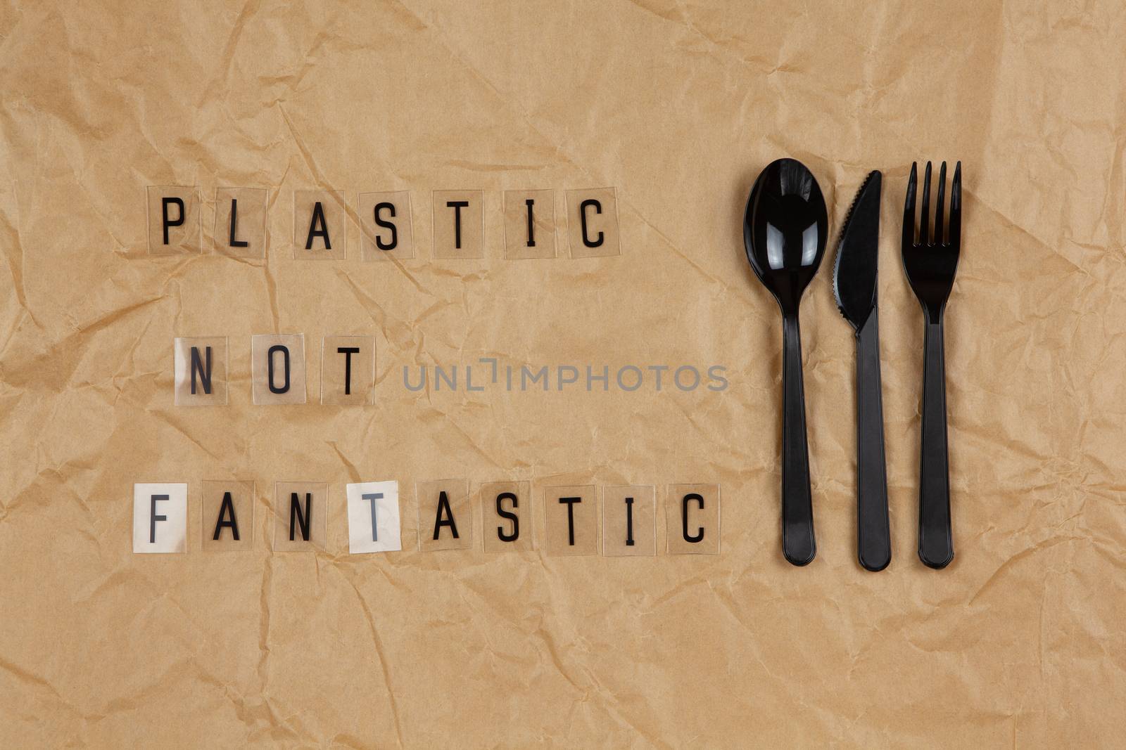 Disposable black appliances fork, spoon, knife, phrase from letters on transparent base Plastic not fantastic on brown crumpled craft paper. Eco, zero waste concept. Flat lay, top view. Horizontal.