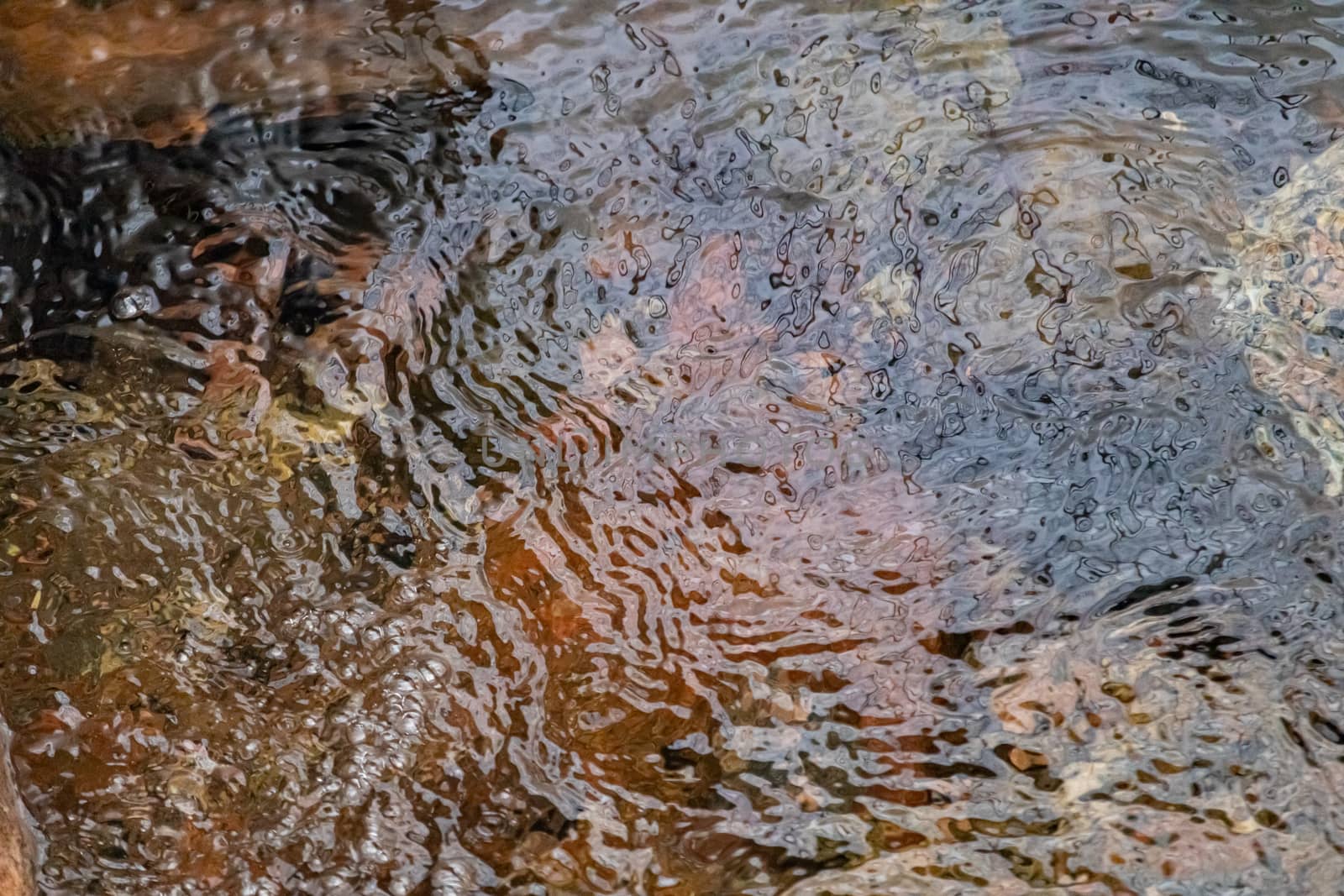 Water ripples in a small stream as it flows over brown and reddish rocks below. The transparent surface shows the shape and texture of the water's surface.