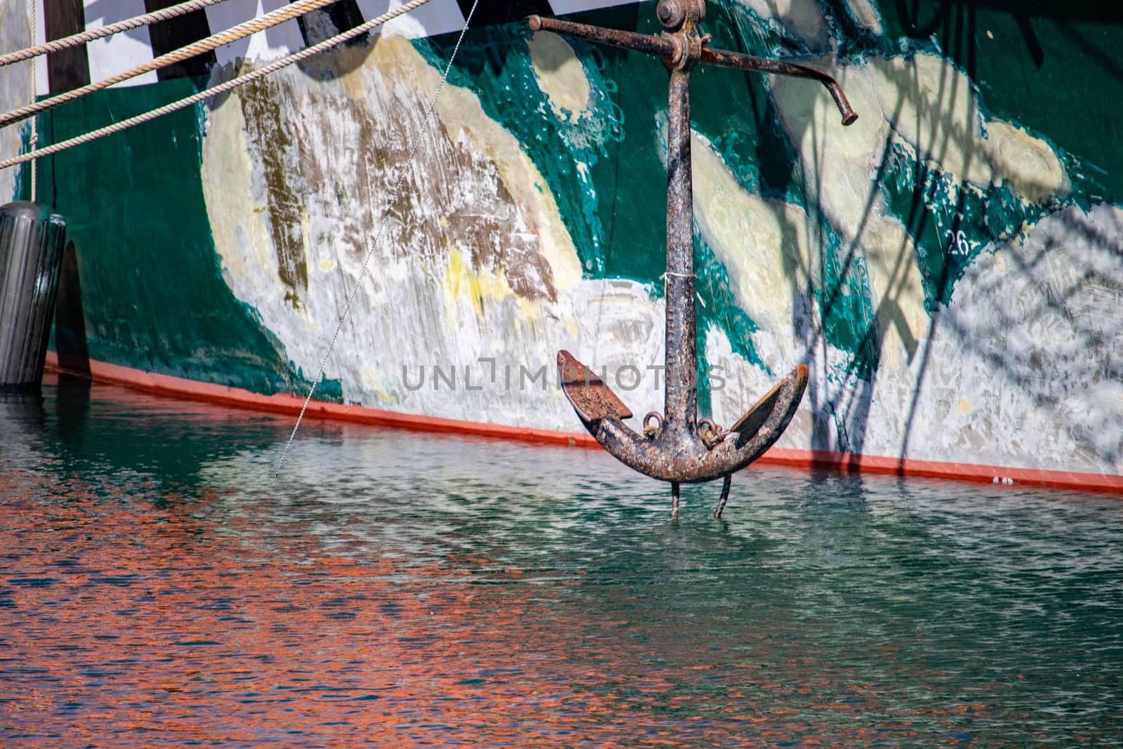 The anchor of a grungy boat is seen hanging above rippling water before the boat's hull.