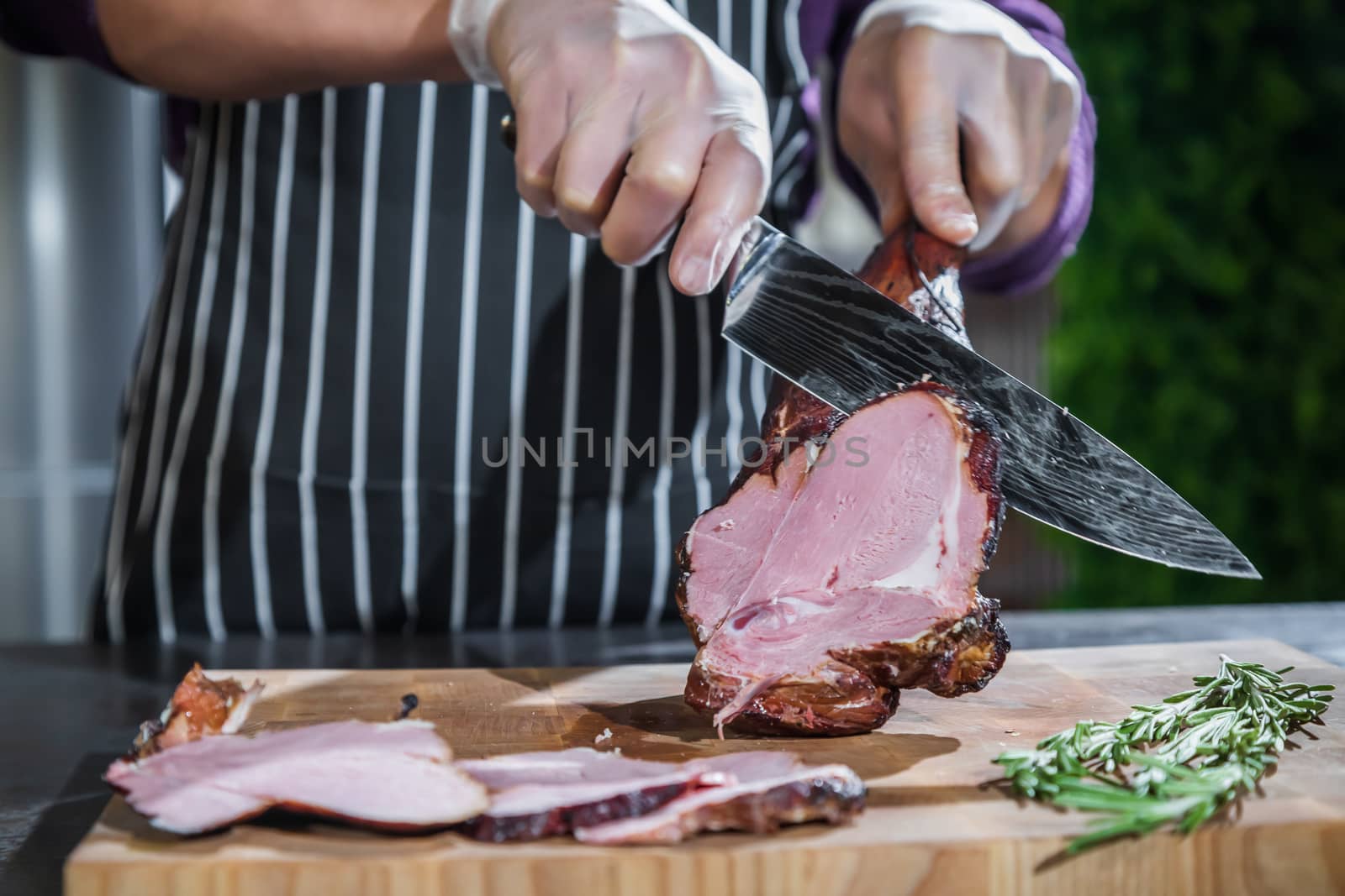 A cook cuts a smoked lamb leg into slices on a wooden cutting board with a knife.