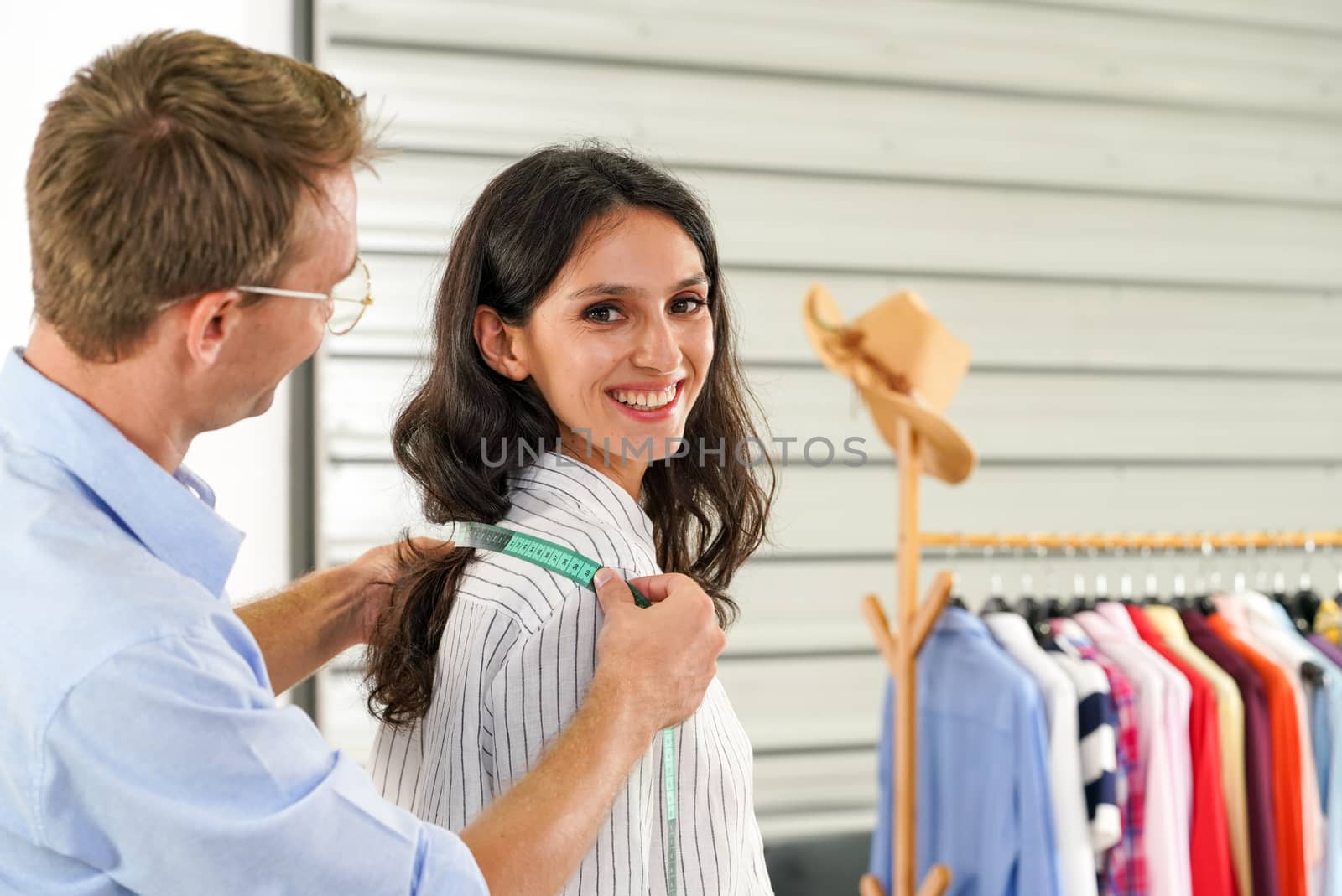 Fashion designer room, man and woman working in home base cloth design business. A good men's tailor is measuring the shoulder size of a young customer. Customer smiled while ordering a new outfit.