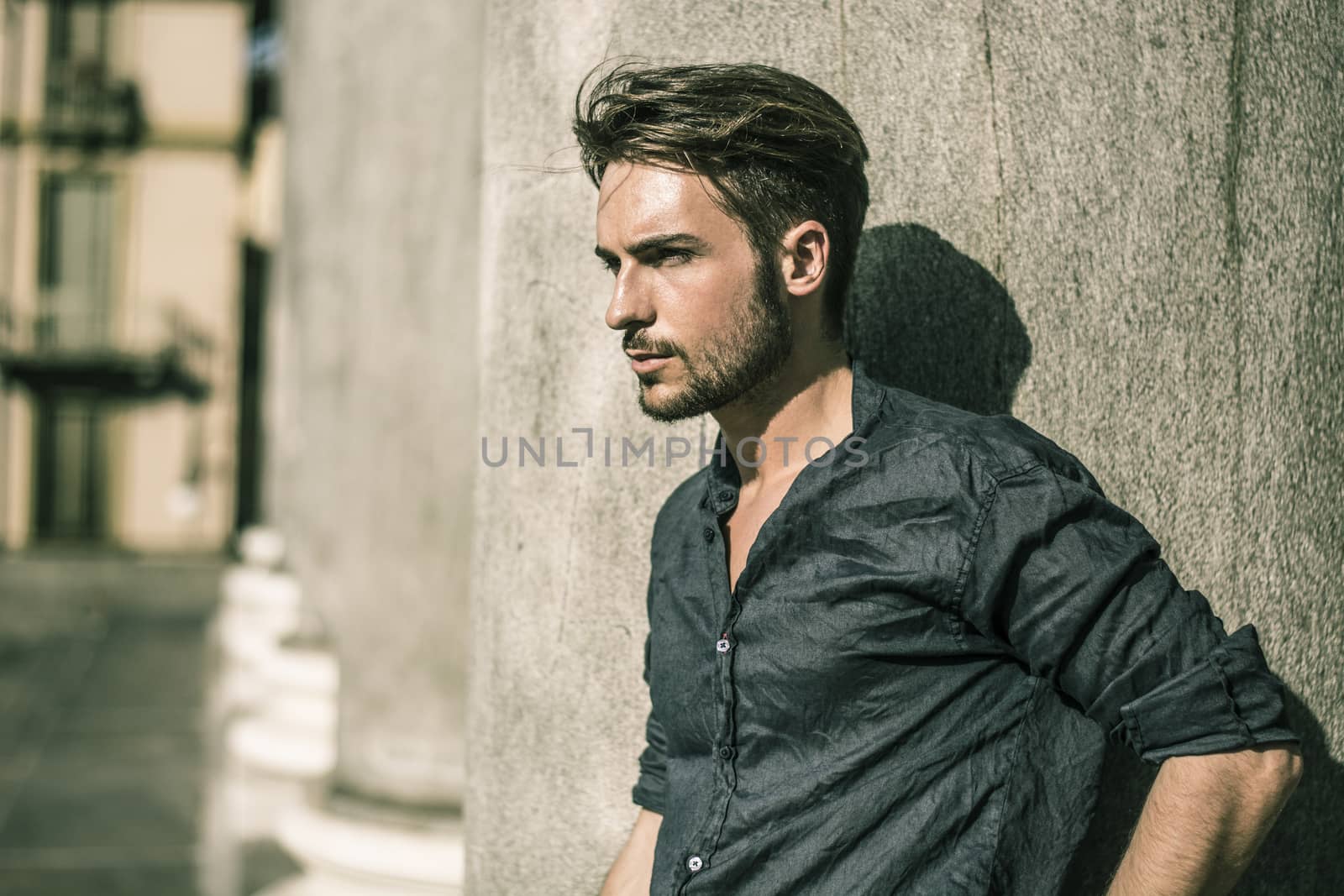 One handsome young man in urban setting in summer day, wearing blue shirt, leaning against stone column or pillar