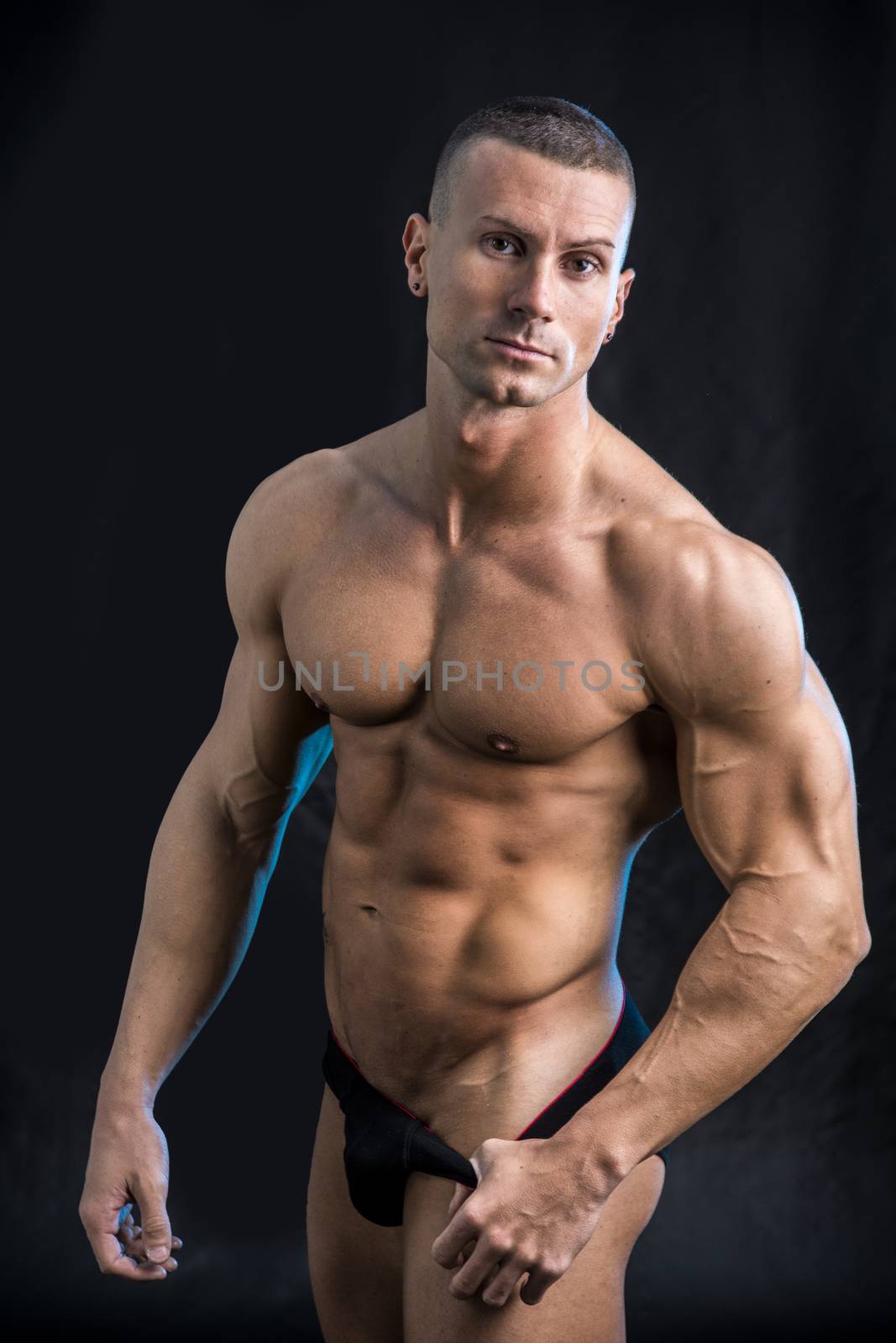 Handsome confident shirtless bodybuilder standing in underwear, looking at camera, on black background