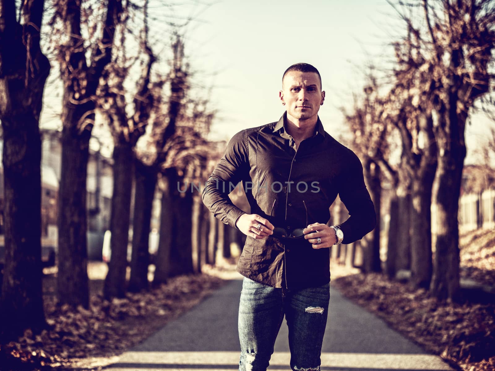 One handsome young man in urban setting in European city walking along street with trees, wearing black jacket and jeans