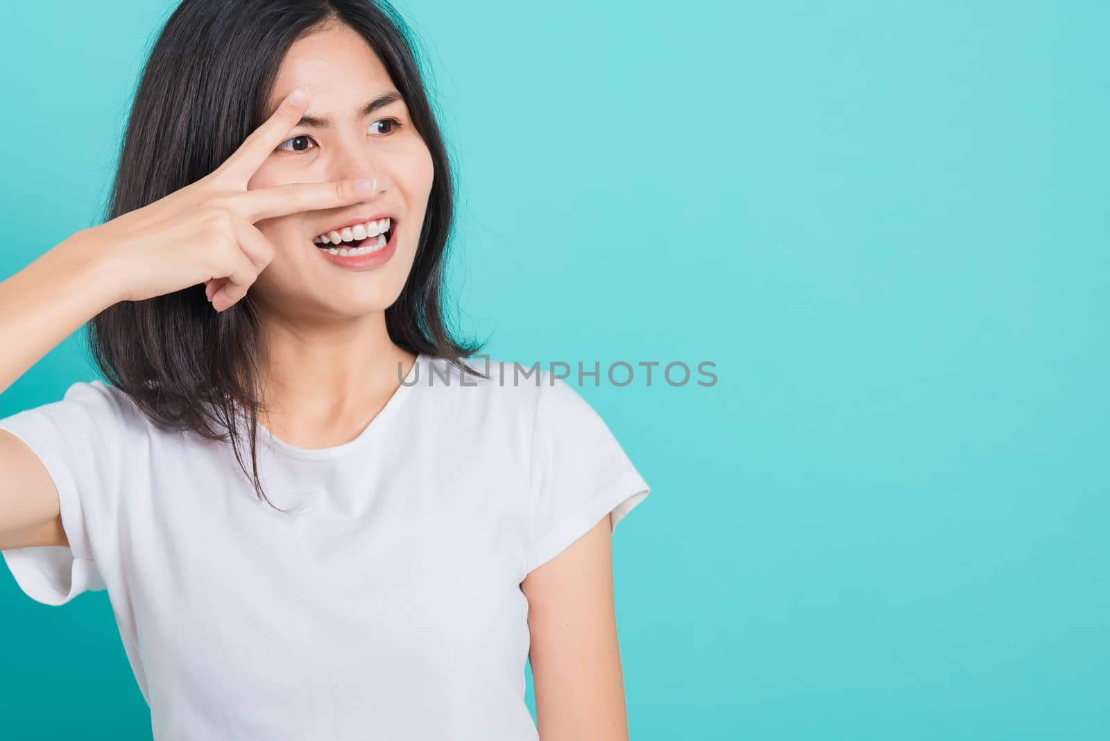 Portrait Asian beautiful happy young woman smile white teeth wear white t-shirt standing showing two finger making v-sign near the eye, on a blue background with copy space