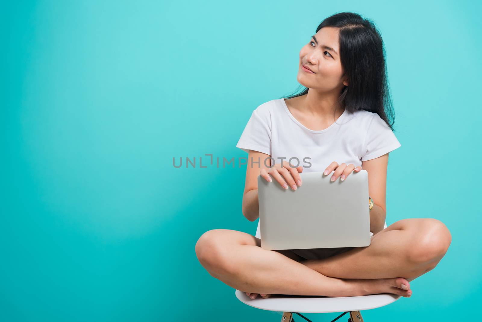 Portrait happy Asian beautiful young woman smile with teeth sitting on chair wear white t-shirt, She holding and using a laptop computer, studio shot on blue background with copy space for text