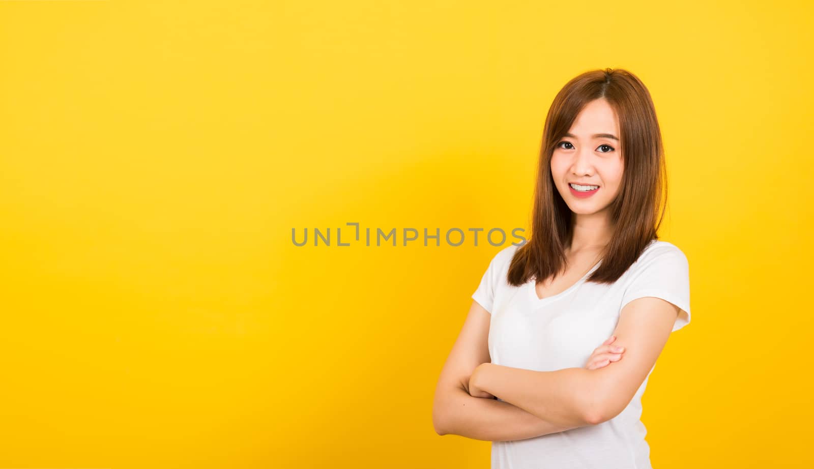 Asian happy portrait beautiful cute young woman teen standing wear t-shirt smile her confidence with crossed arms looking to camera isolated, studio shot on yellow background with banner copy space