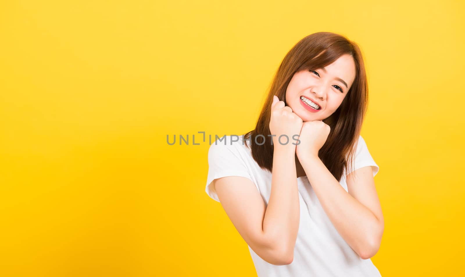 Asian happy portrait beautiful cute young woman teen stand wear t-shirt happy expression fist pressed together under chin looking to camera isolated, studio shot on yellow background with copy space