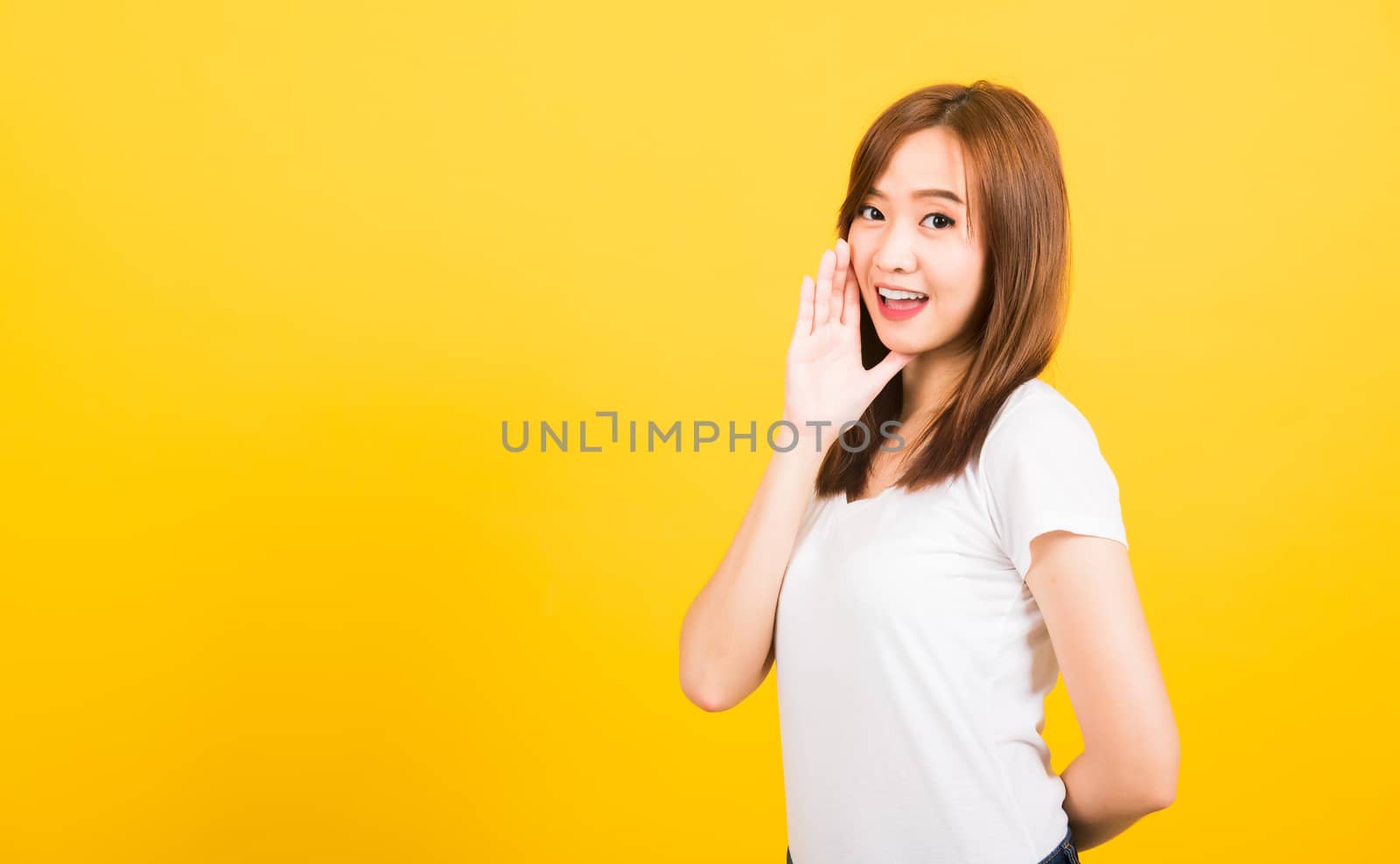 Asian happy portrait beautiful cute young woman teen standing wear t-shirt hand on mouth talking whispering secret rumor looking to camera isolated, studio shot on yellow background with copy space