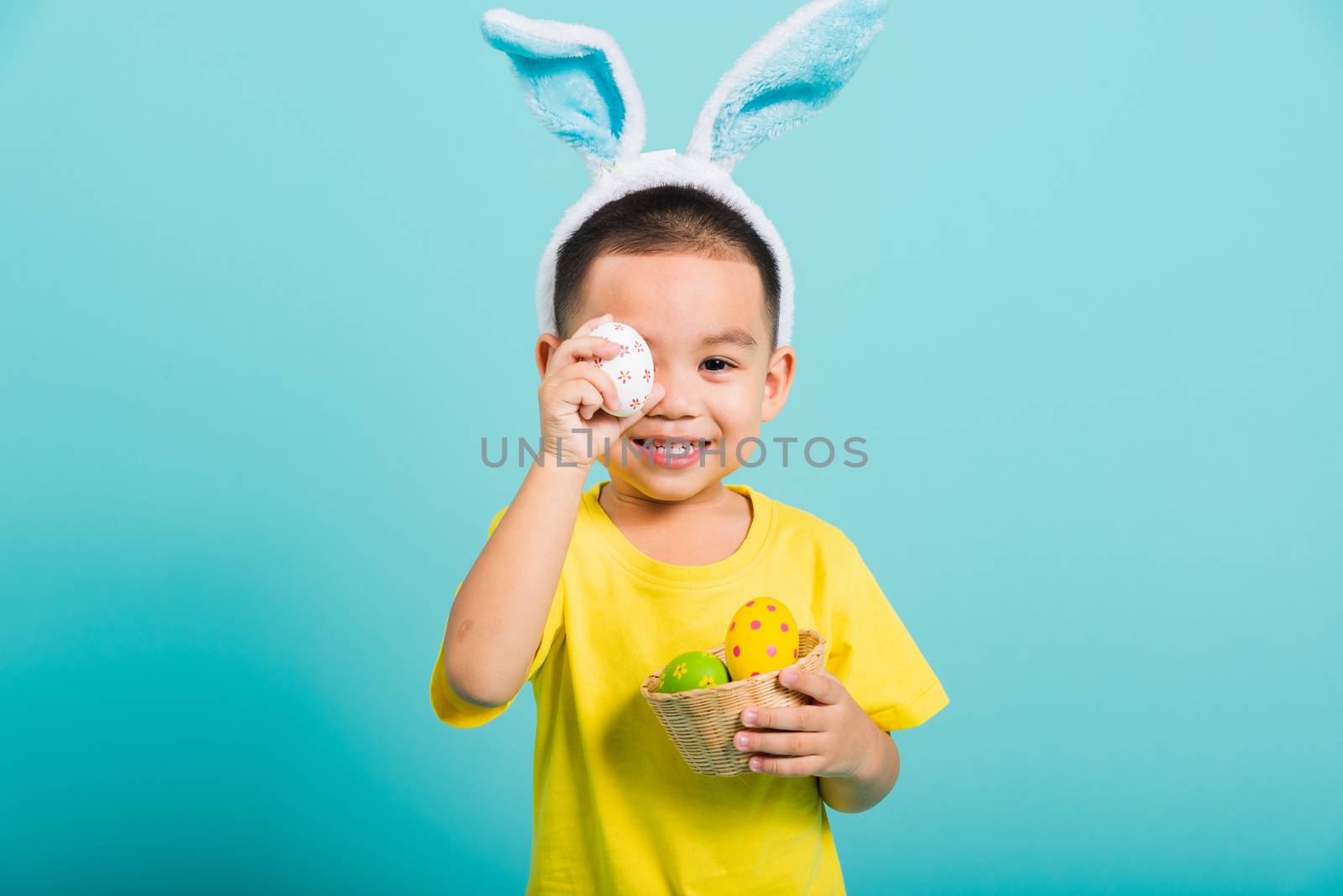 Asian cute little child boy smile beaming wearing bunny ears and a yellow T-shirt, standing to holds colored easter eggs instead of eyes on blue background with copy space for text