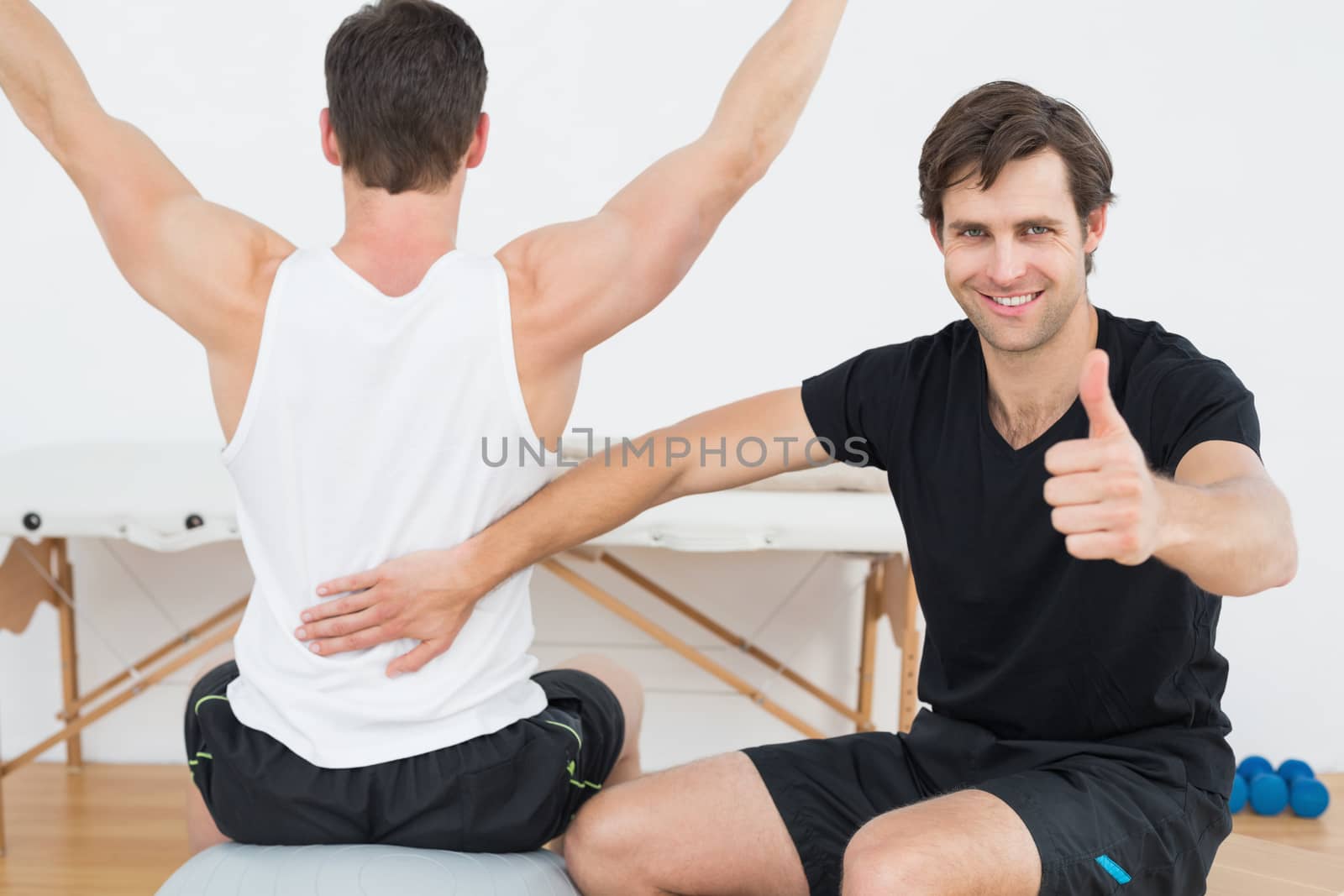 Therapist gestures thumbs up besides man on yoga ball by Wavebreakmedia