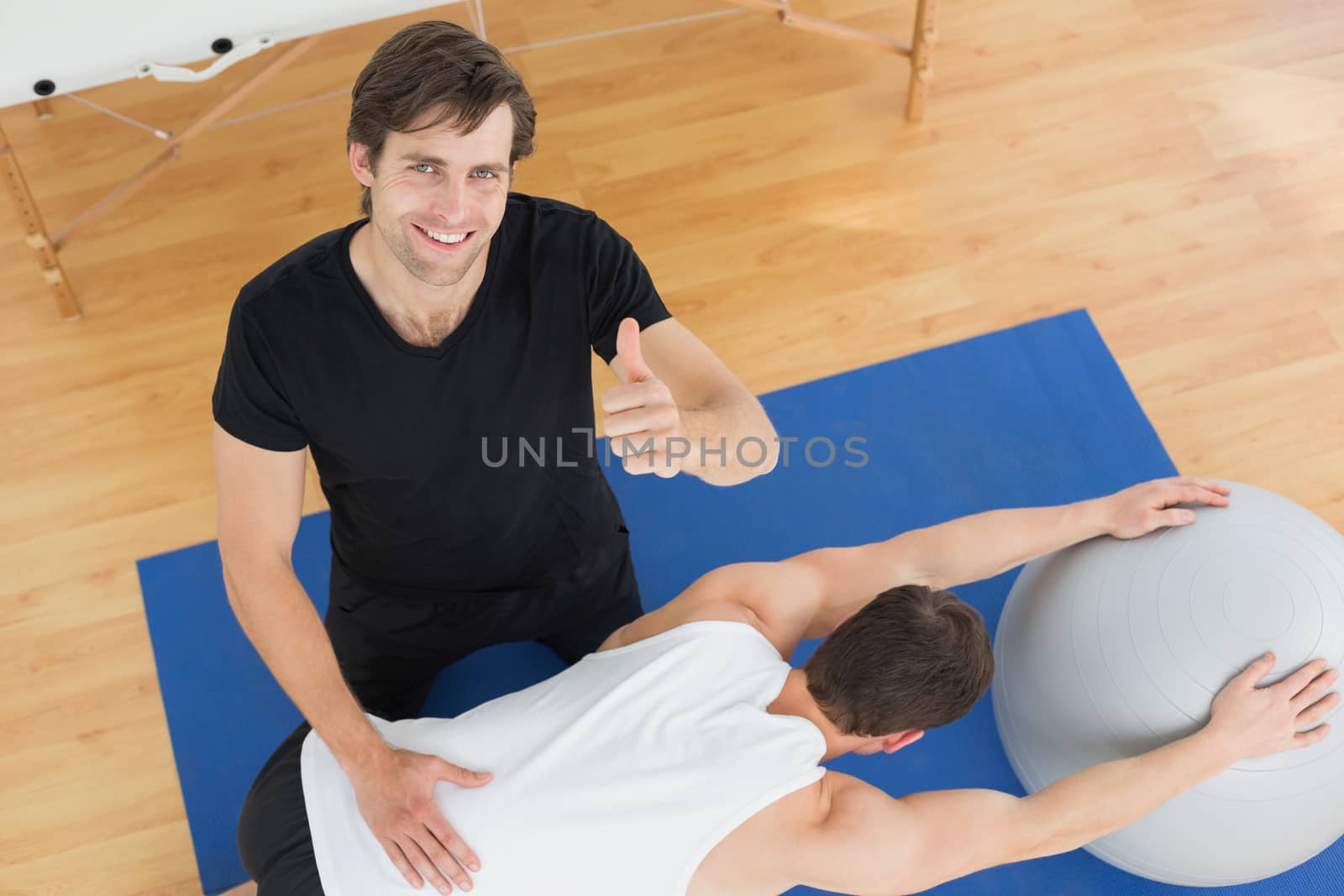 High angle view of a physical therapist gesturing thumbs up by young man with yoga ball in the gym at hospital