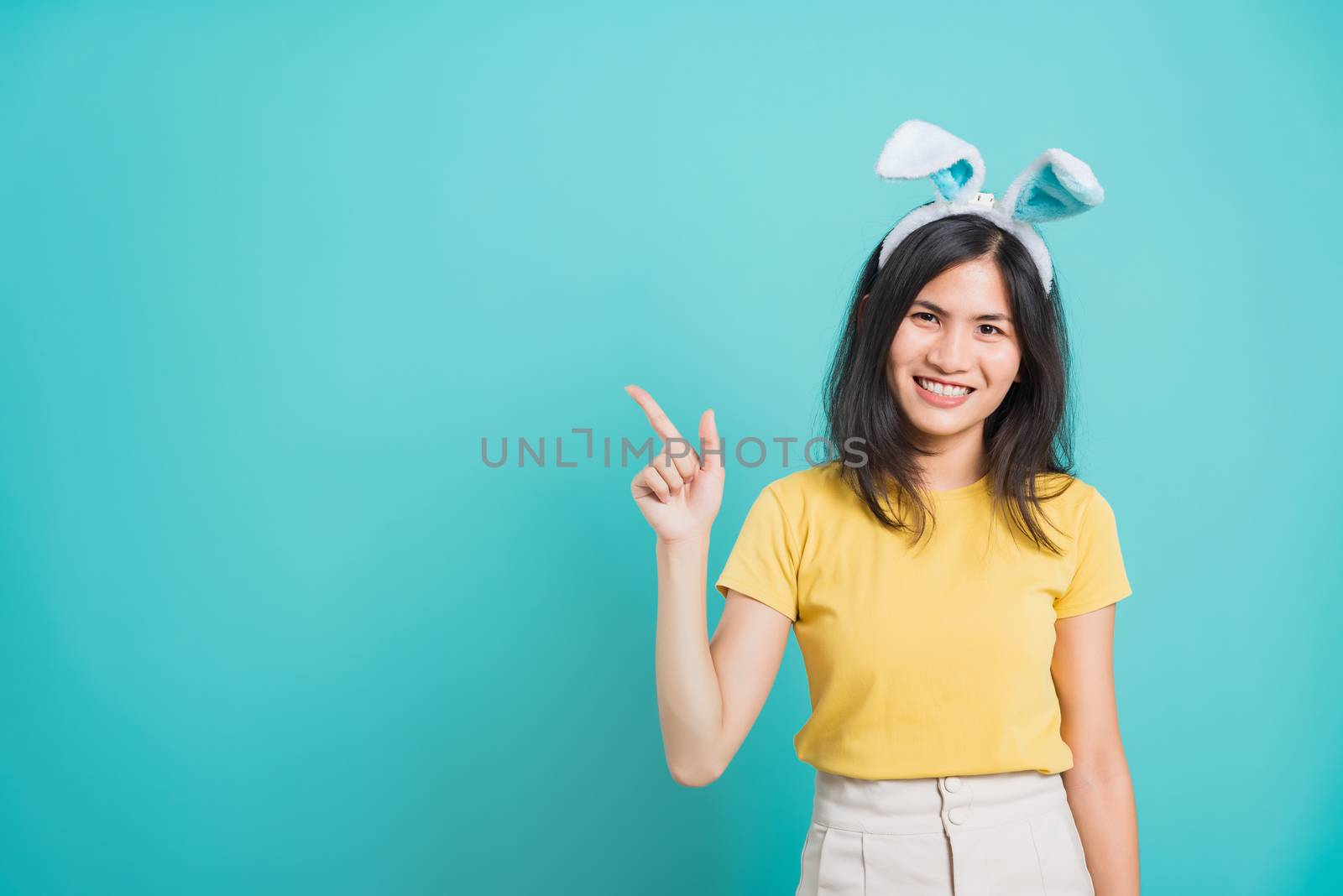 Portrait Asian beautiful happy young woman smile white teeth wear yellow t-shirt standing with bunny ears pointing to space her looking camera, on blue background with copy space