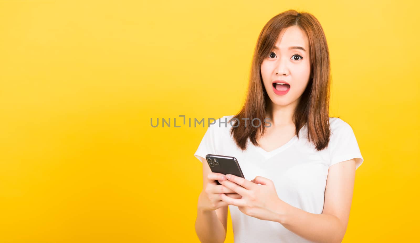 Asian happy portrait beautiful cute young woman teen smiling standing wear t-shirt her surprised excited with smart mobile phone looking camera isolated, studio shot yellow background with copy space