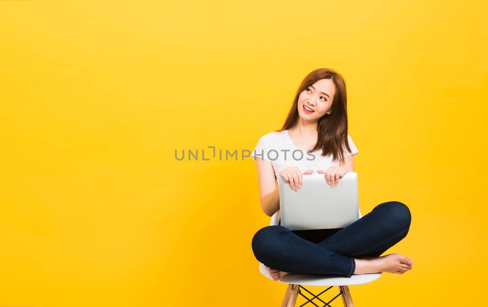 Asian happy portrait beautiful cute young woman teen smiling sitting crossed legs on a chair with laptop computer looking to side isolated, studio shot on yellow background with copy space