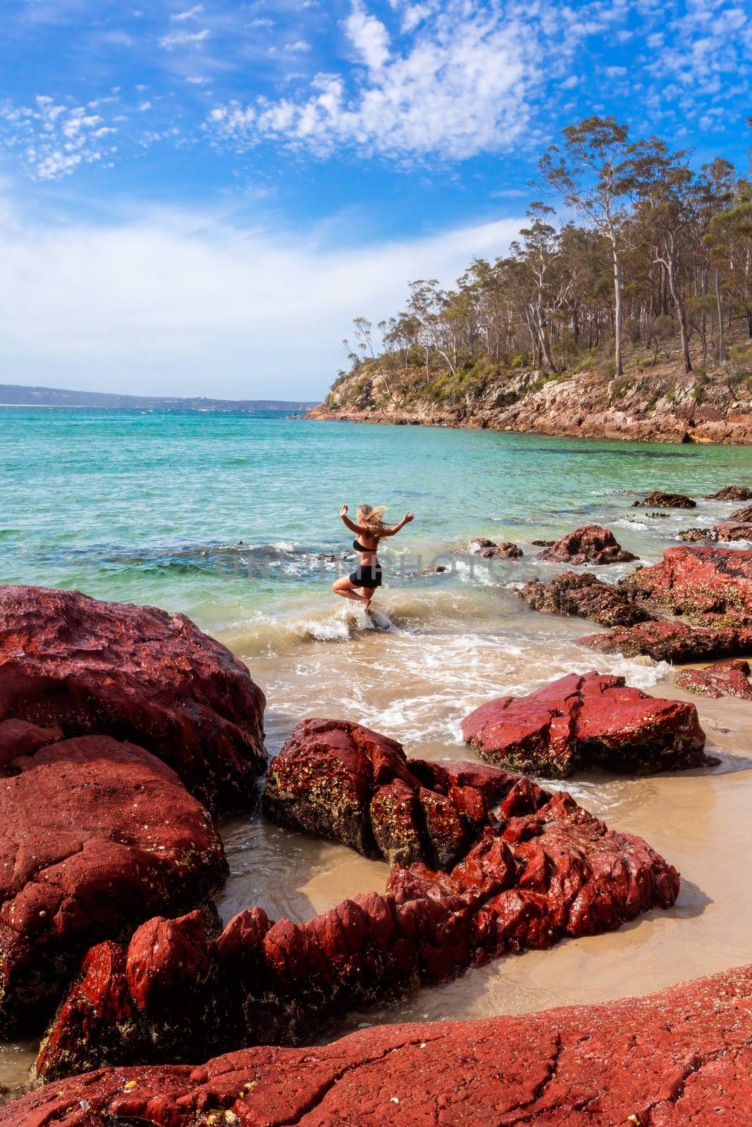 A woman playing in beach paradise, running into the aqua blue waters of the ocean.  A scattering of rich red rocks in the foreground