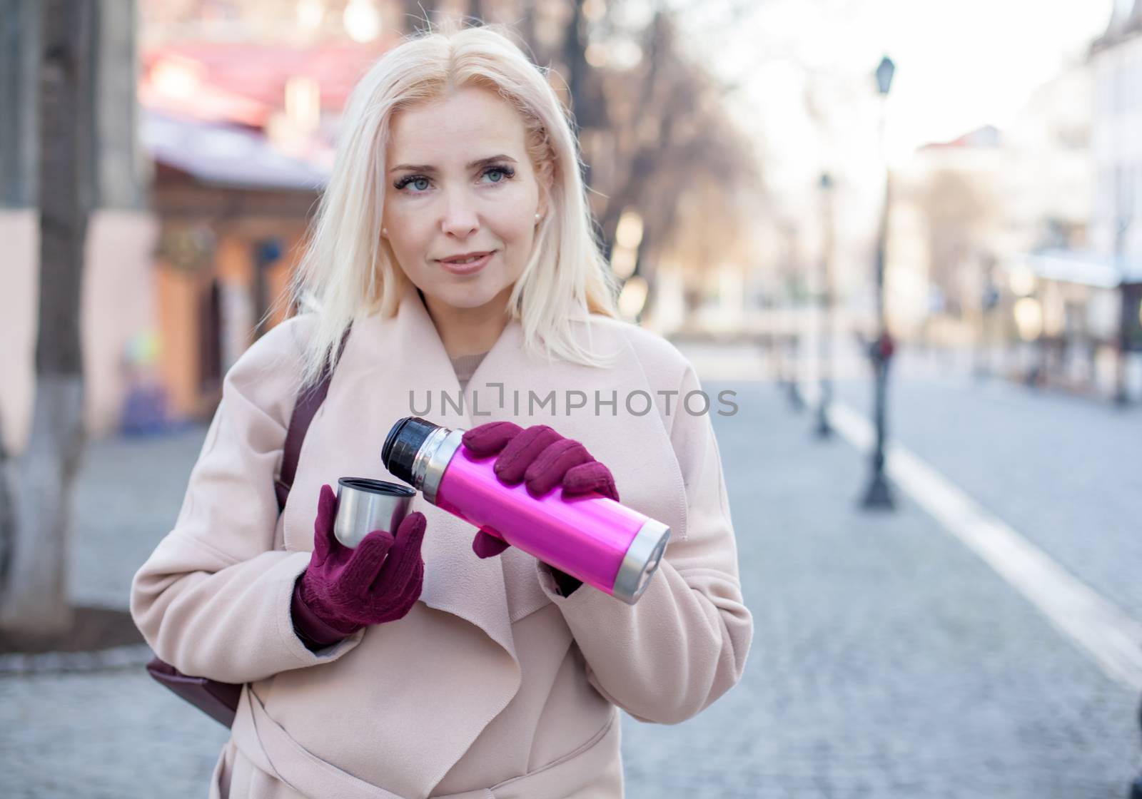 Outdoor of gorgeous woman with blond hair in elegant coat and gloves walking in street with thermos of tea