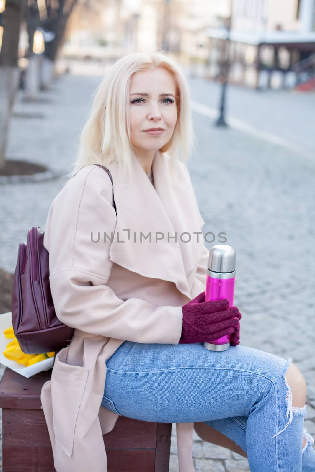 Outdoor of gorgeous woman with blond hair in elegant coat and gloves sitting in street with thermos of hot tea