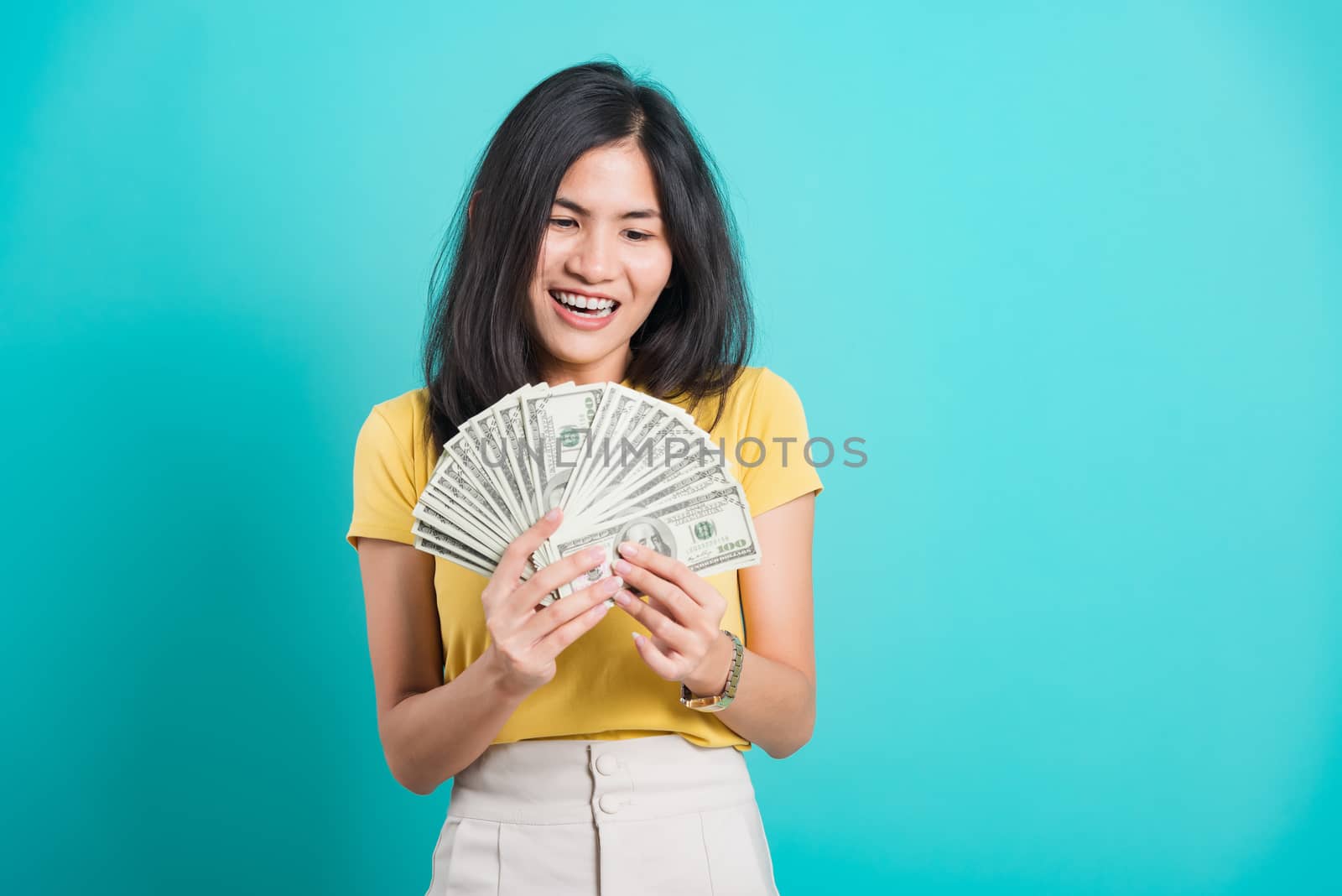 Asian happy portrait beautiful young woman standing wear t-shirt smiling holding money fan banknotes 100 dollar bills and looking to money isolated on blue background with copy space for text