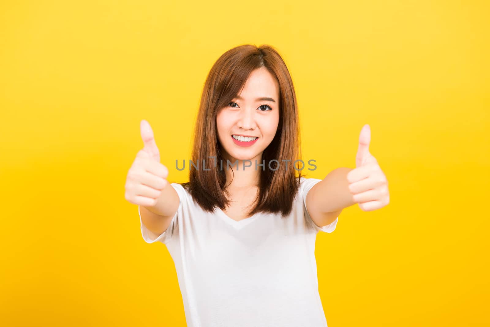 Asian happy portrait beautiful cute young woman teen standing wear t-shirt showing gesturing finger thumb up looking to camera isolated, studio shot on yellow background with copy space