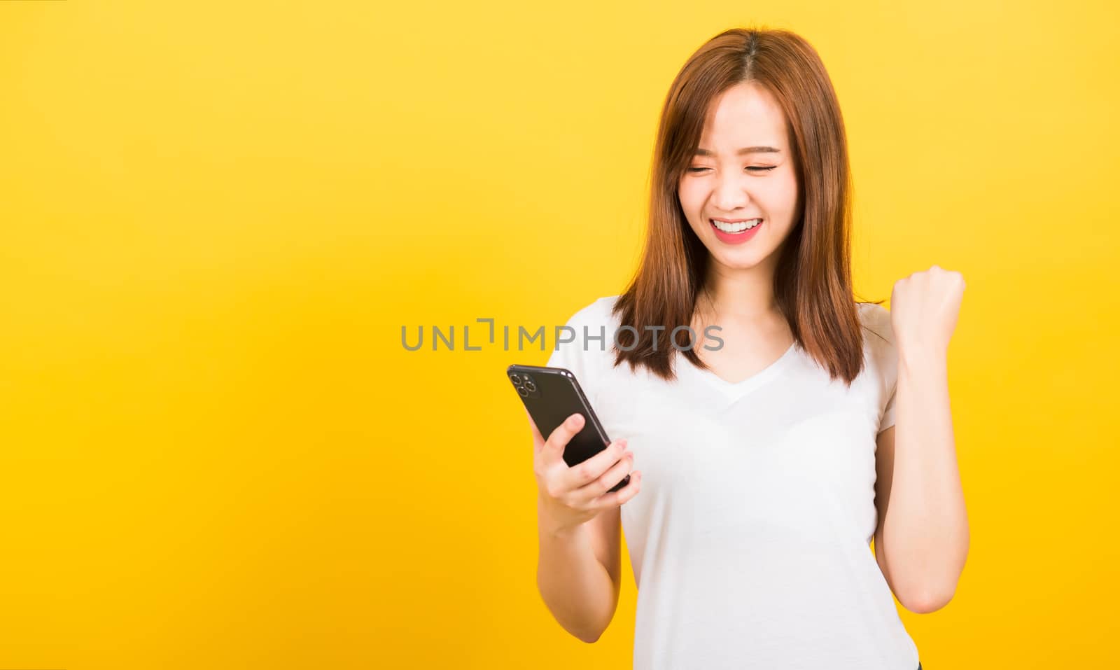 Asian happy portrait beautiful cute young woman teen smiling standing wear t-shirt celebrating win with smart mobile phone looking the phone isolated, studio shot on yellow background with copy space