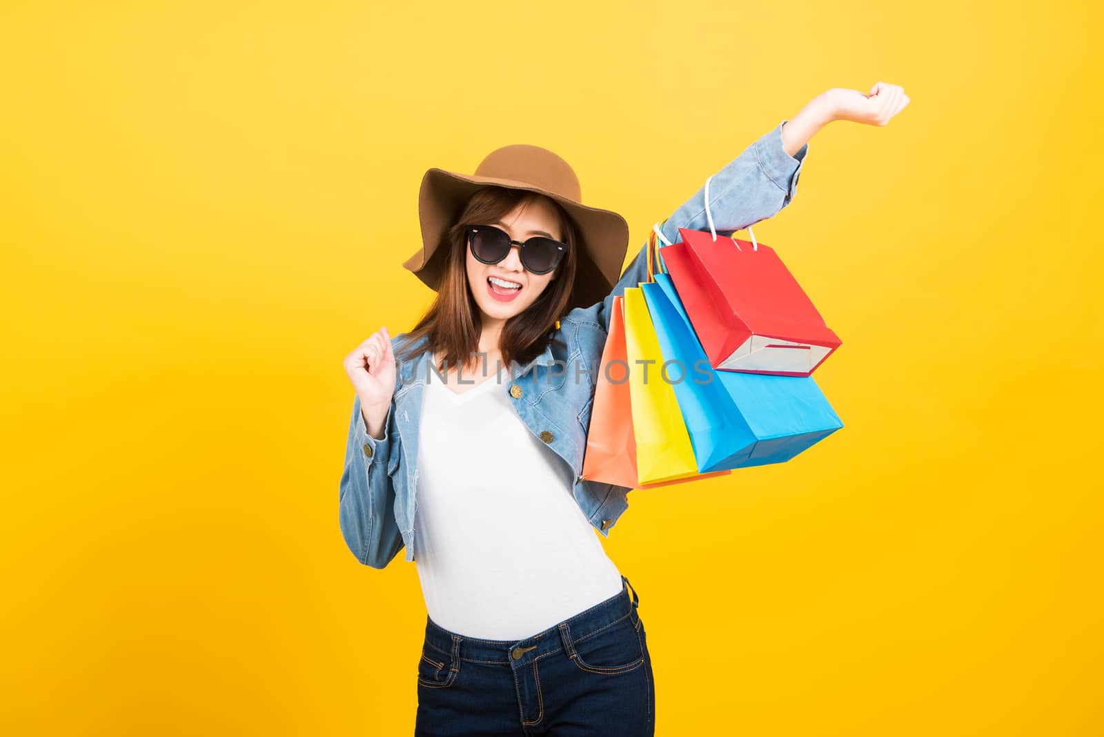 Asian happy portrait beautiful cute young woman teen smiling standing with sunglasses excited holding shopping bags multi color looking camera isolated, studio shot yellow background with copy space