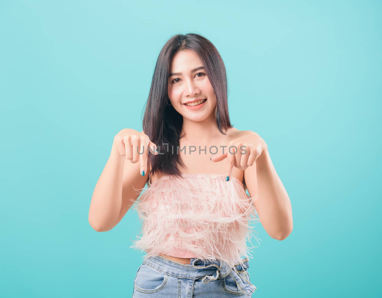 Asian happy portrait beautiful young woman standing smiling pointing finger down and looking to camera isolated on blue background with copy space for text